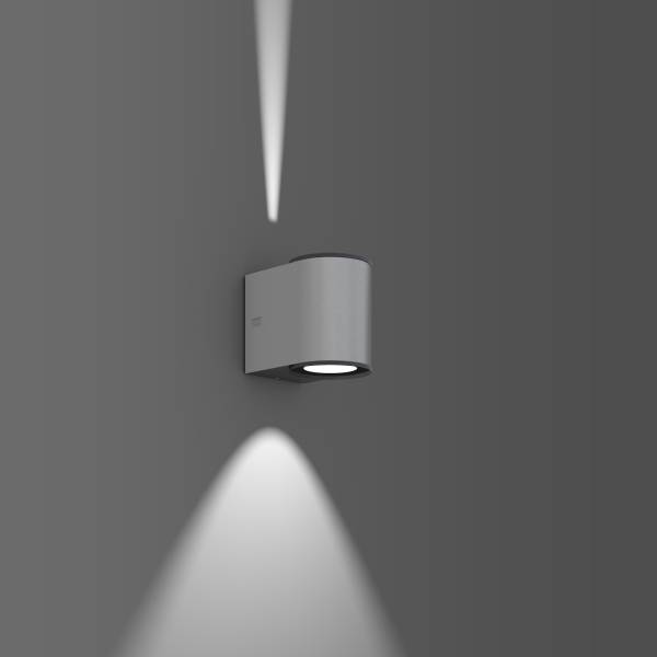 Moderní nástěnná lampa v šedé barvě, která vyzařuje světlo jak nahoru, tak dolů, namontovaná na šedé zdi.