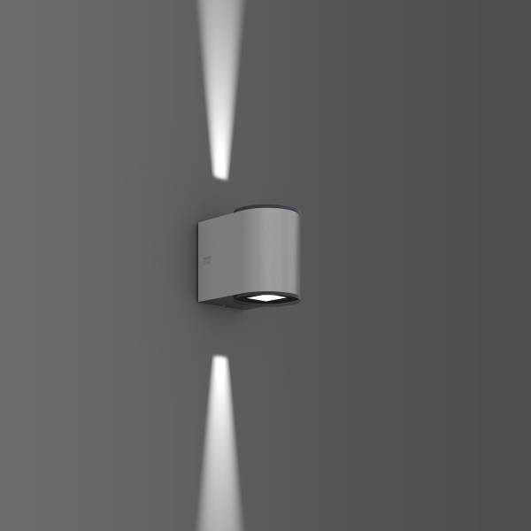 Nástěnná lampa na šedé stěně, svítí nahoru a dolů, vytváří zaostřené světelné kužely.