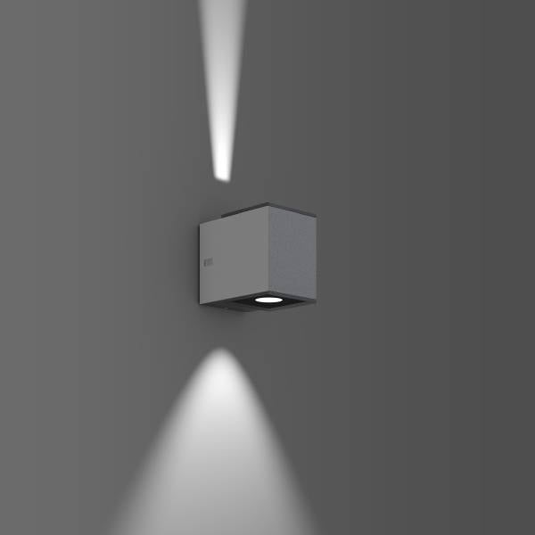 Nástěnná lampa s kubickým designem osvětluje šedou zeď světelnými paprsky směřujícími nahoru a dolů.
