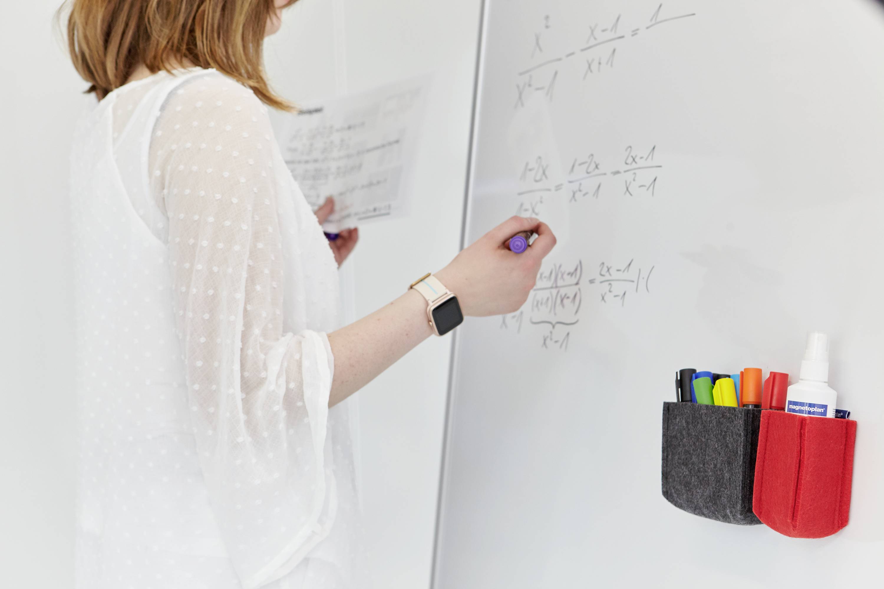 Jedna osoba píše matematické vzorce na whiteboard. Vedle jsou barevné permanentní fixy v držáku.