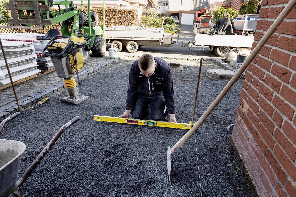 Muž klečí na staveništi a používá vodováhu k vyrovnání povrchu. V pozadí jsou stavební zařízení a cihlový dům.