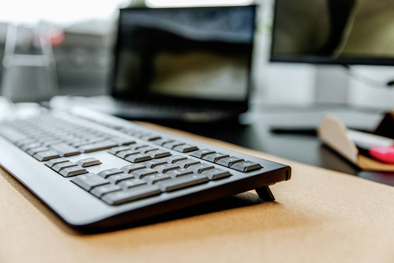 Psací stůl s rozostřeným monitorem počítače v pozadí, v popředí moderní černá klávesnice, zobrazující pracovní prostředí.