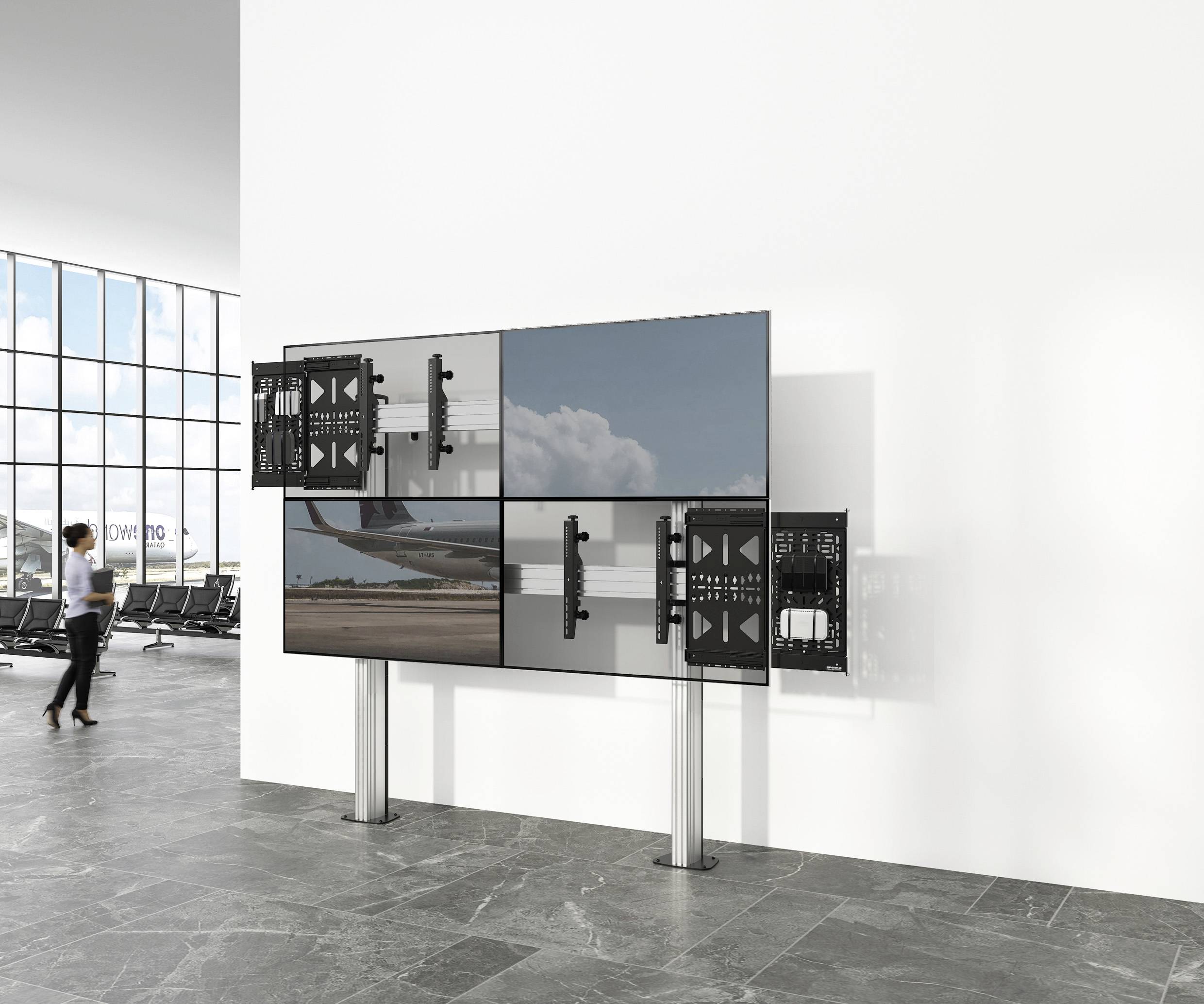 Čekací prostor letiště s velkým šestidílným displejem. Chodec prochází kolem prosklené stěny.