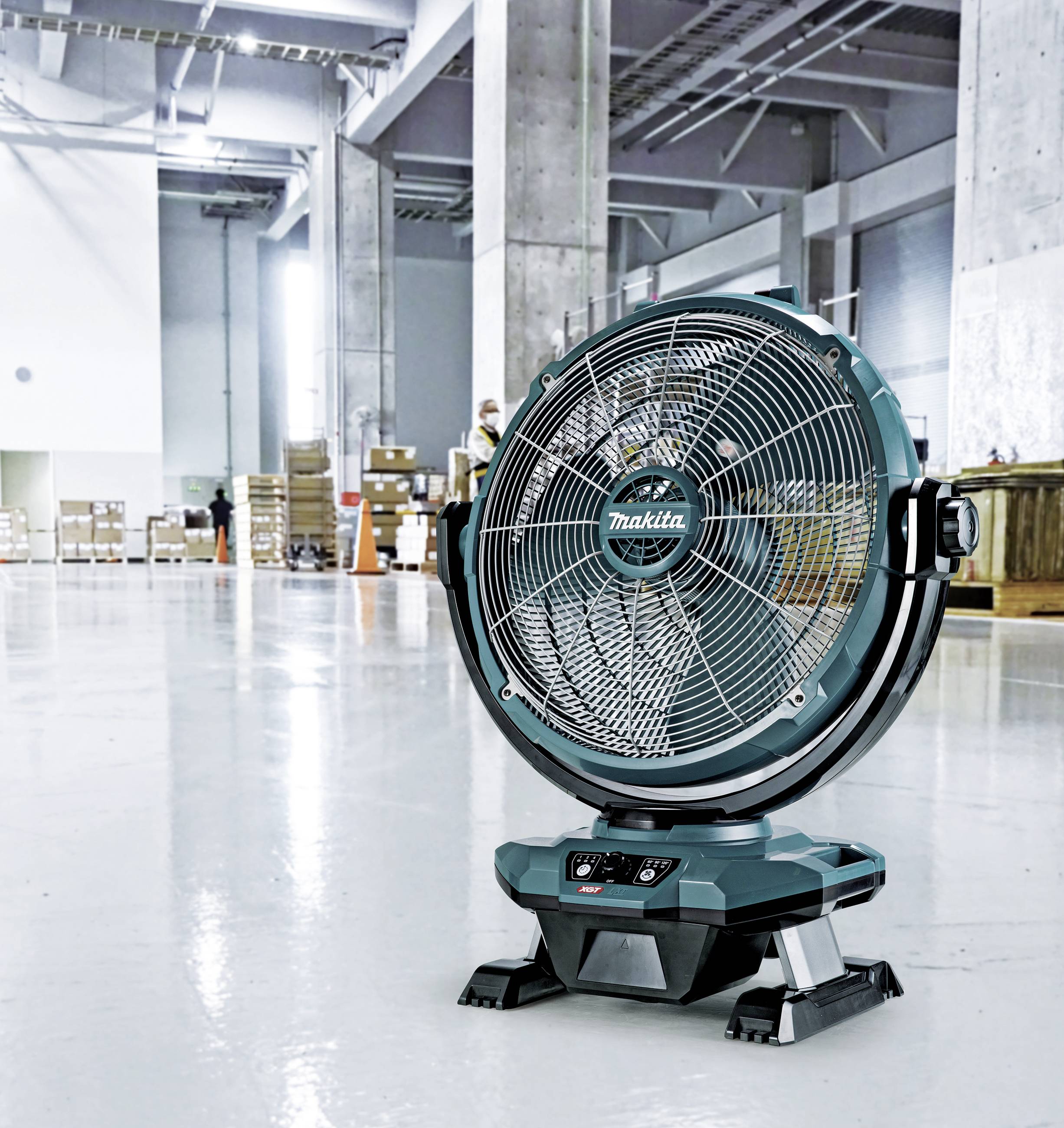 Zelený průmyslový ventilátor stojí na lesklé tovární podlaze s regály a krabicemi v pozadí, osvětlen horními světlíky.