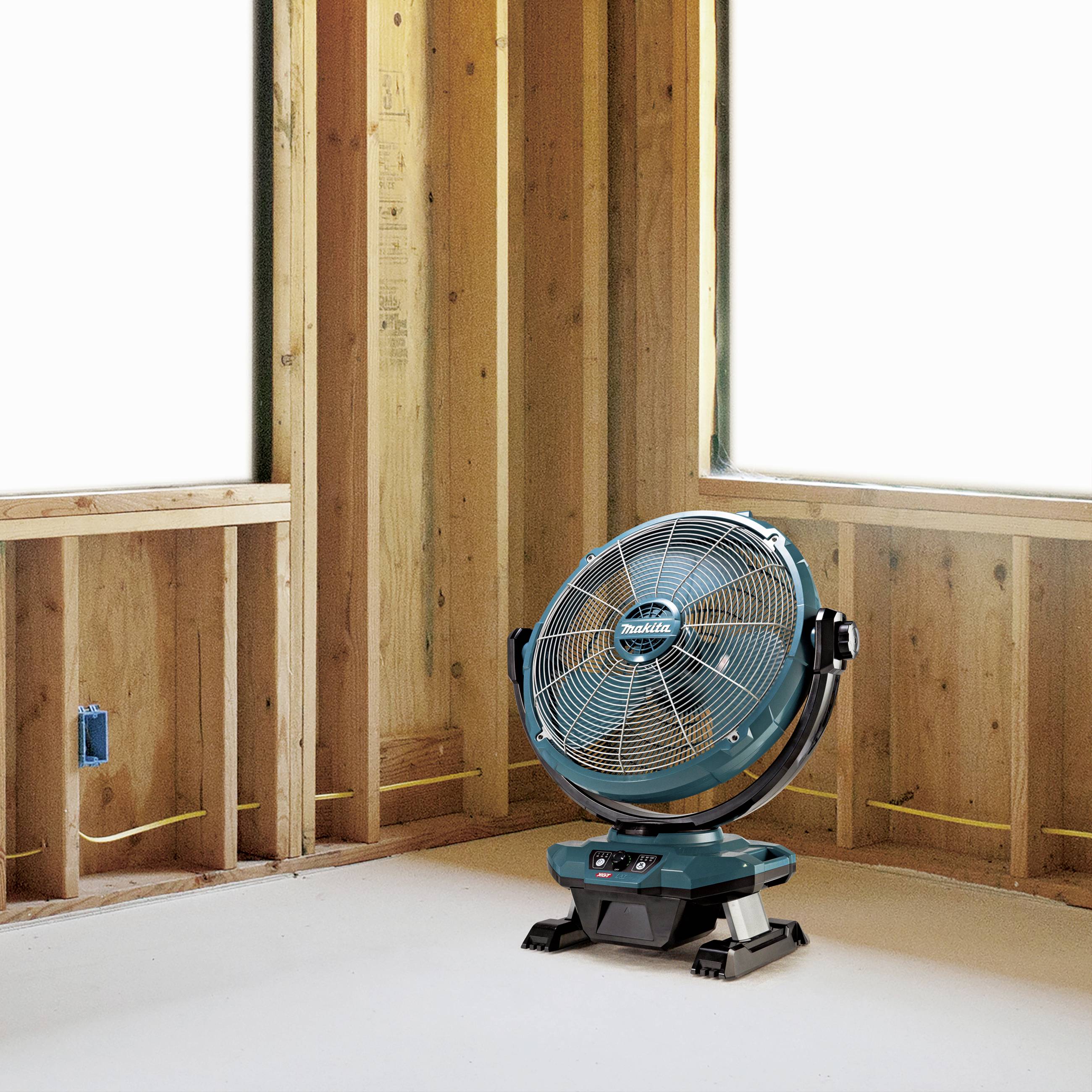 Konstrukční zóna s viditelnými dřevěnými rámy, kabely a zásuvkou. Na podlaze stojí modrý ventilátor, který vhání vzduch do místnosti.