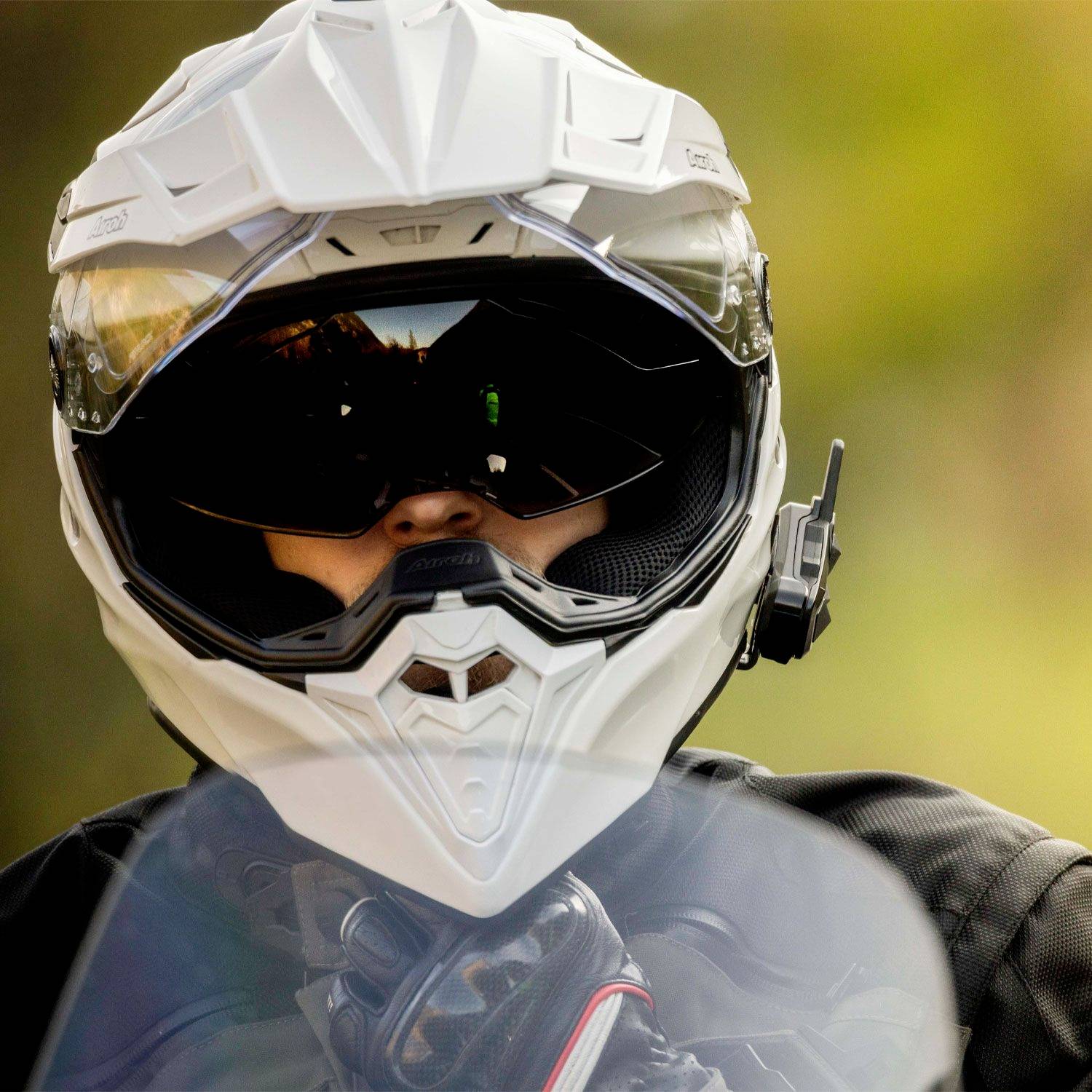 Osoba má na sobě bílou motocyklovou helmu a černé ochranné brýle, v pozadí je rozmazaná zeleň a modř.