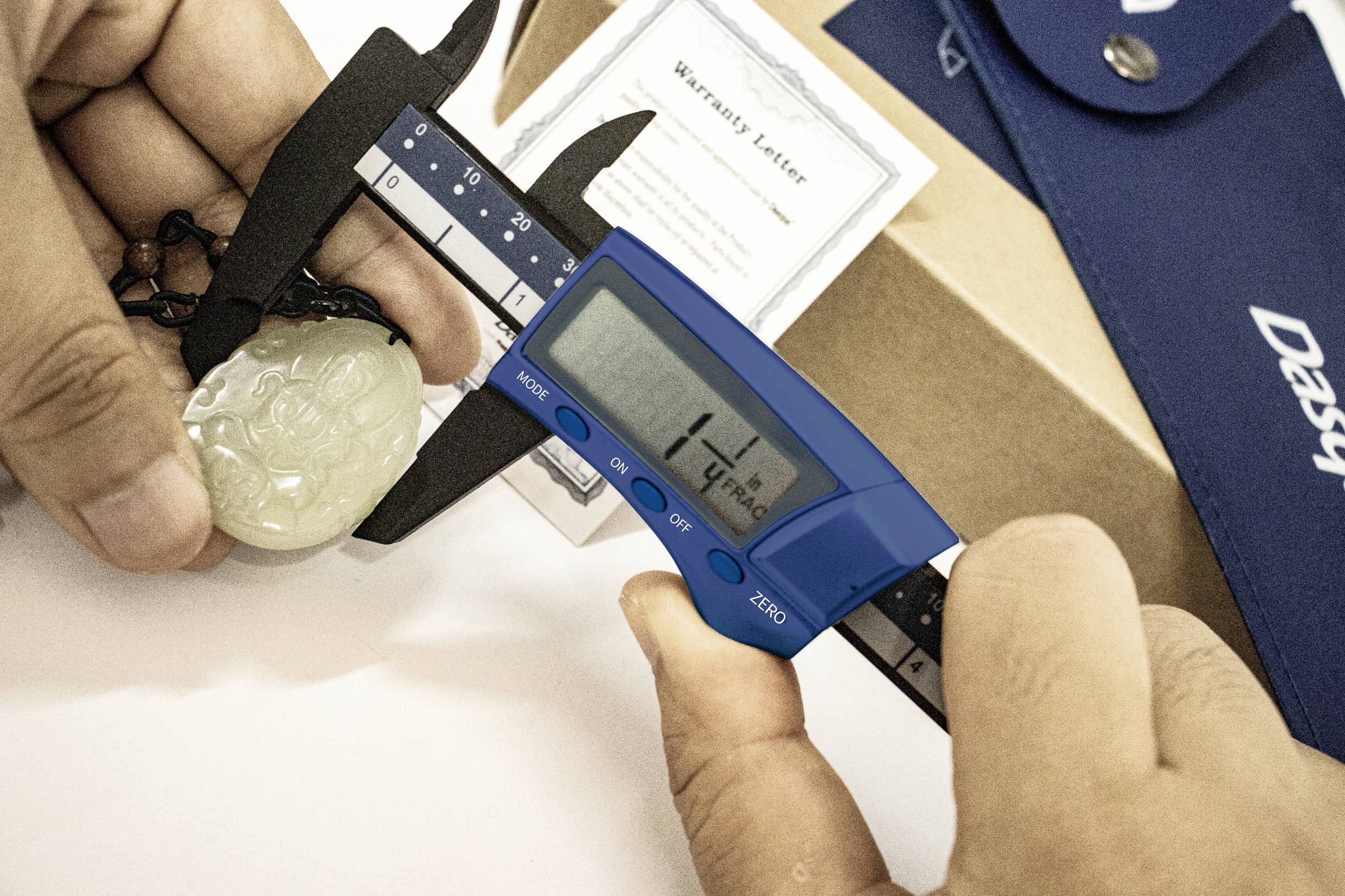 Jedna ruka drží digitální posuvné měřidlo, které měří drahokam. Na displeji je zobrazeno '47.8mm'. V pozadí je vidět certifikát a obal.