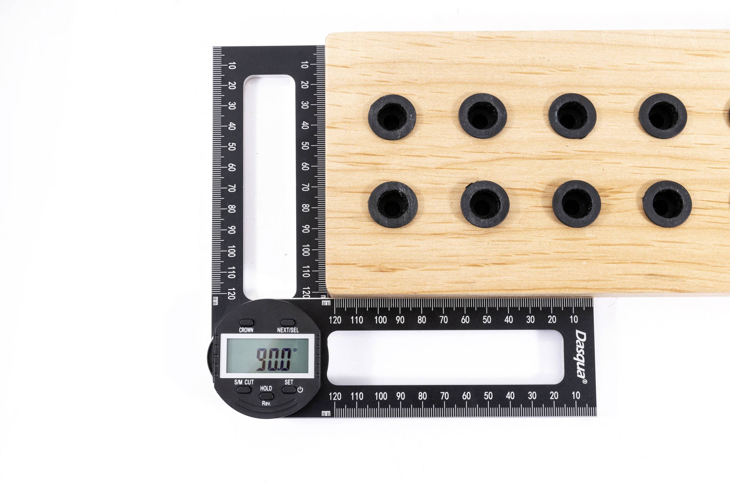 Digitální posuvné měřítko ukazuje 90,0 mm. Měří vzdálenost mezi dvěma body na dřevěné desce s černými otvory.