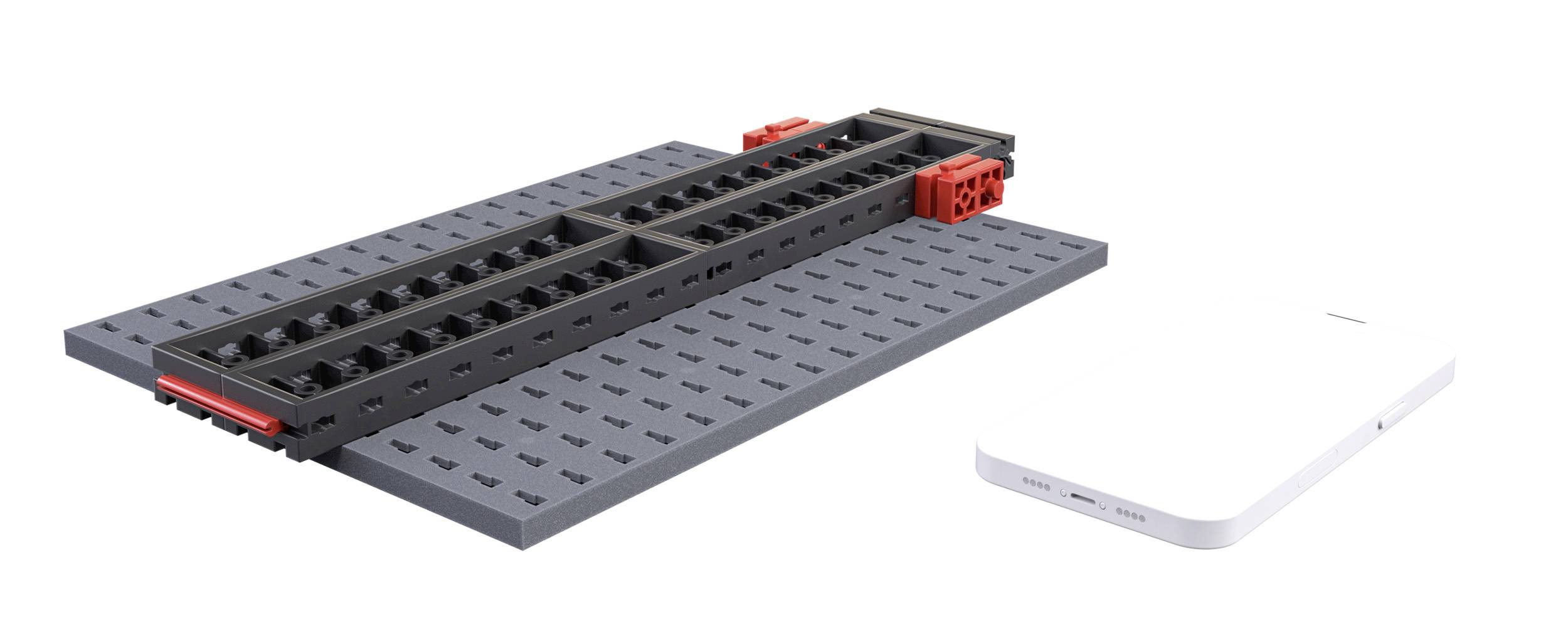 Černý a červený prvek LEGO Technic leží nalevo na šedé desce, vedle něj vpravo bílý obdélníkový objekt.