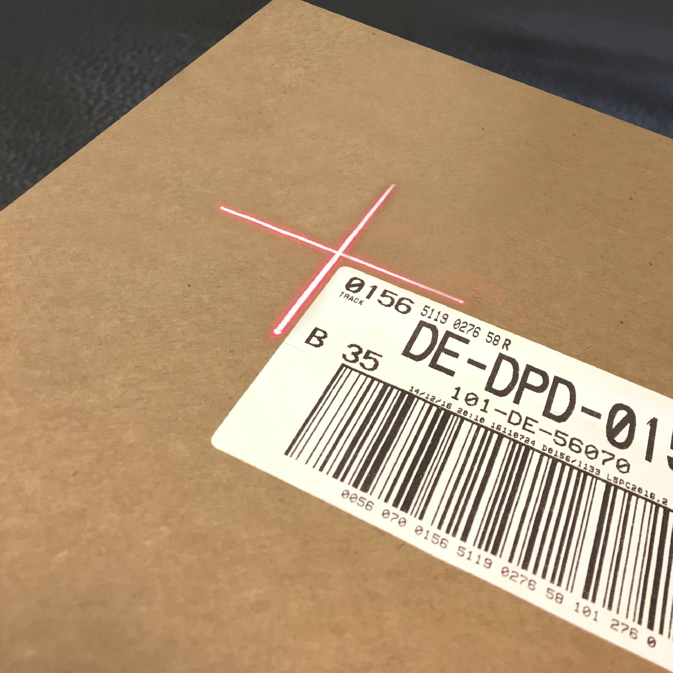 Hnědý balík s naskenovaným čárovým kódem a expedičním štítkem, na kterém je napsáno 'DE-DPD-0156'. Je vidět červený skenovací kříž.