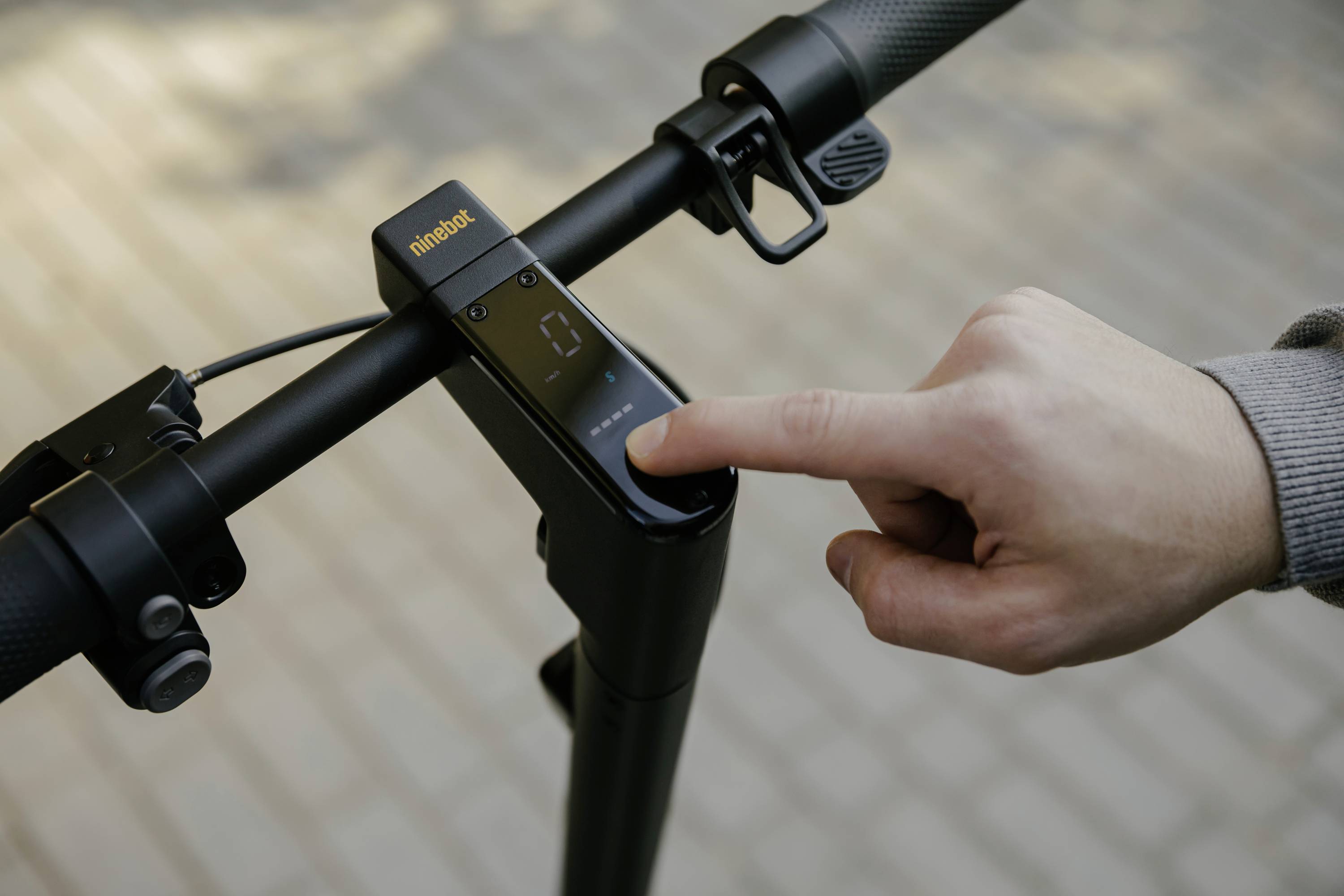Jedna ruka stiskává zapínací tlačítko elektrické koloběžky, jejíž displej na řídítkách ukazuje nulu. Koloběžka stojí na dlážděném povrchu.