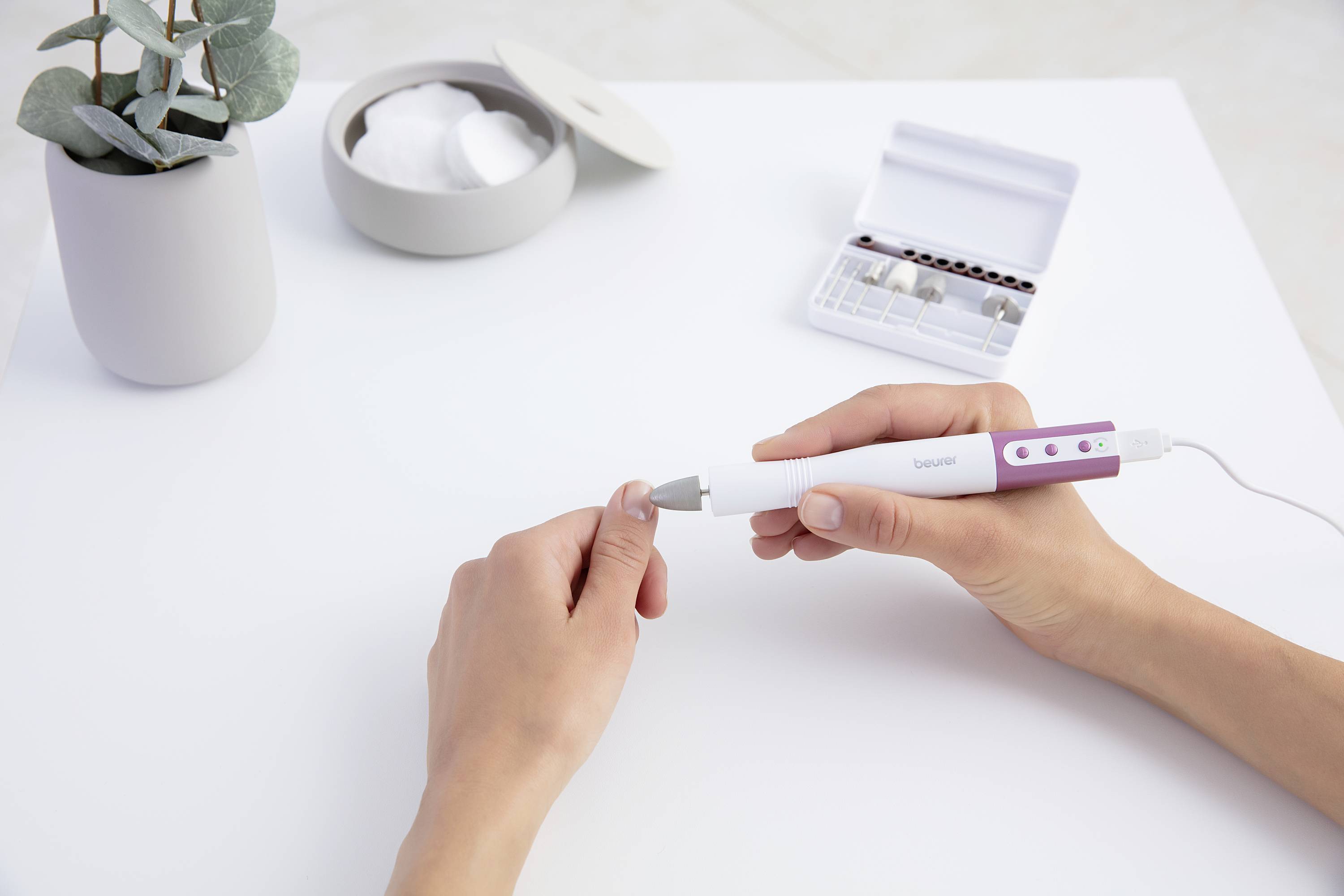 Žena si ošetřuje nehty pomocí elektrického přístroje na péči o nehty. Vedle ní leží otevřená sada nástavců na stole.