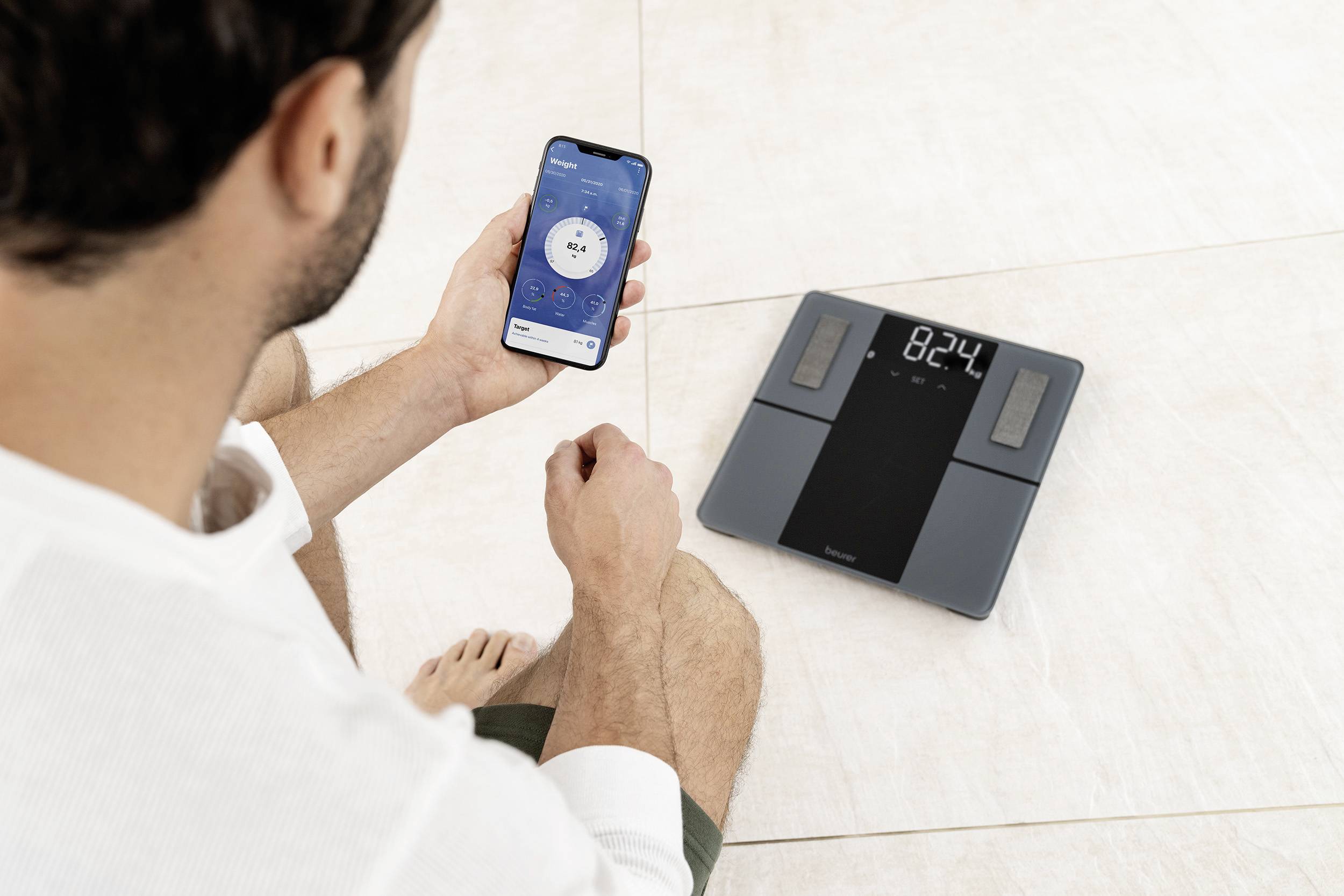 Muž sedí na zemi a sleduje mobilní aplikaci synchronizovanou s digitální váhou. Váha ukazuje 82,4 kg.