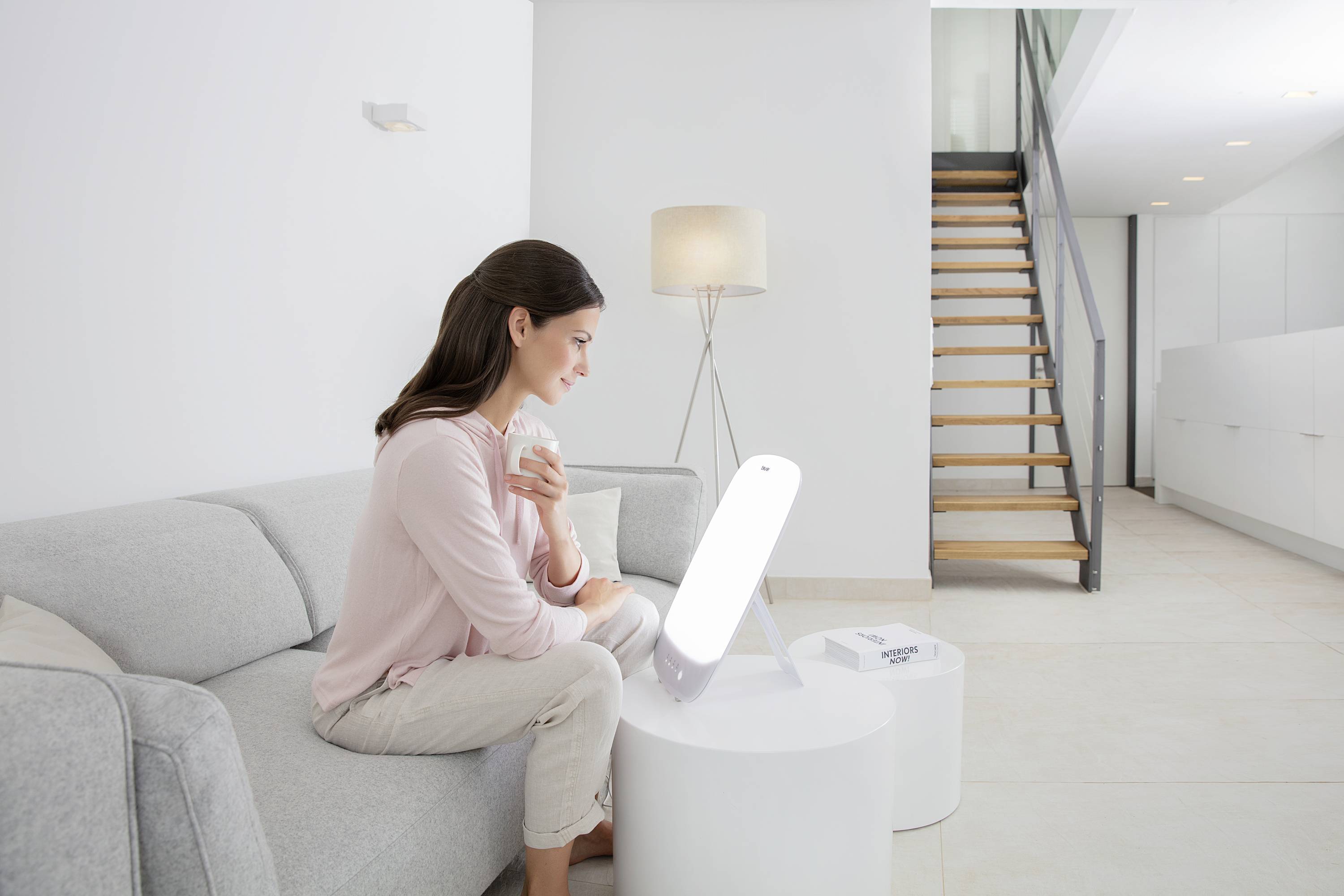 Žena sedí na pohovce ve světlém pokoji, drží šálek a dívá se do světelné terapeutické lampy umístěné na stole.