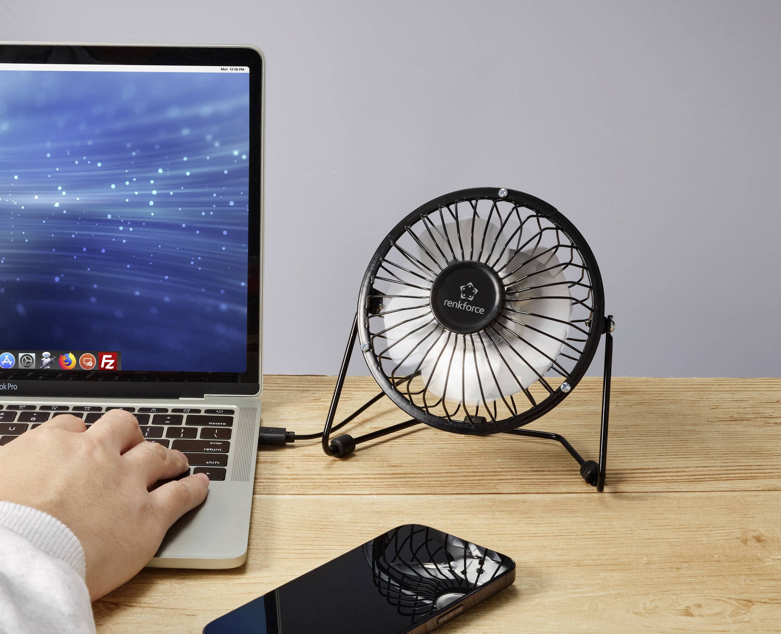 Otevřený laptop vedle malého stolního ventilátoru a smartphonu na dřevěném stole. Ruka píše na klávesnici laptopu.