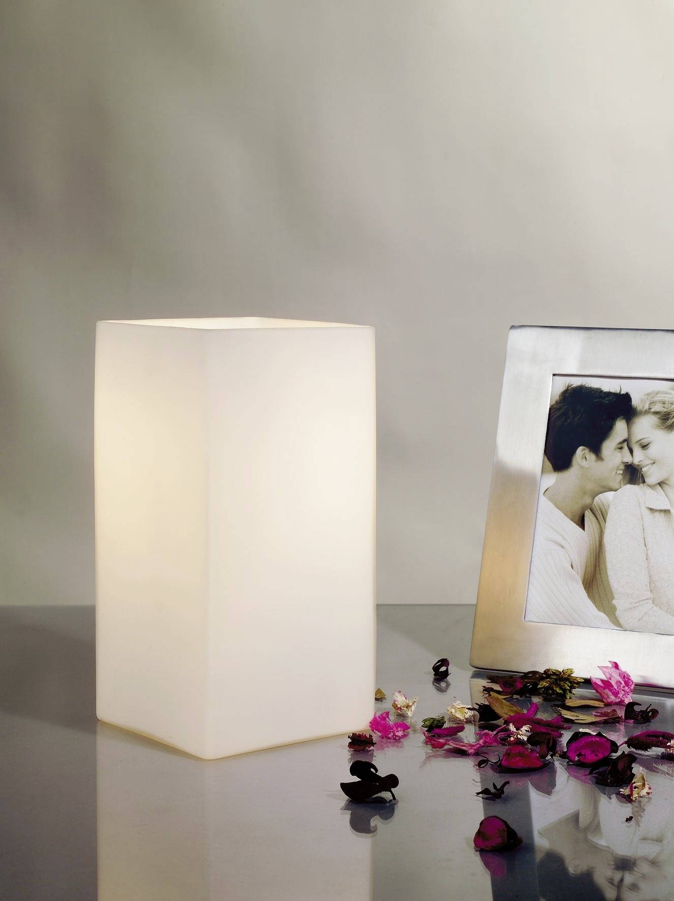 Obdélná stolní lampa vedle rámečku s fotografií páru. Na stole leží růžové okvětní lístky.