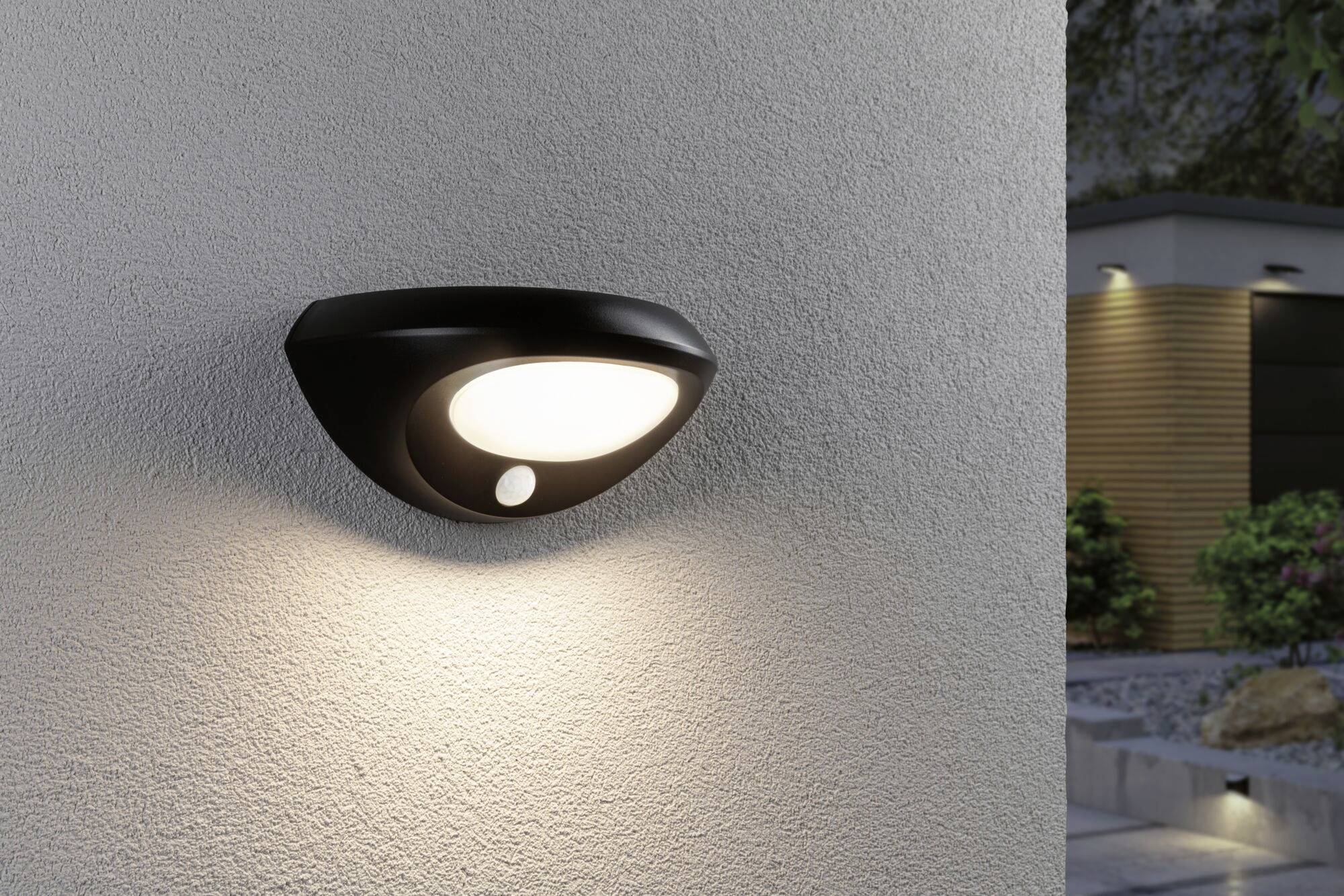 Černobílá venkovní lampa připevněná na stěně svítí v noci. V pozadí rozostřený pohled na zahradu a moderní dům.