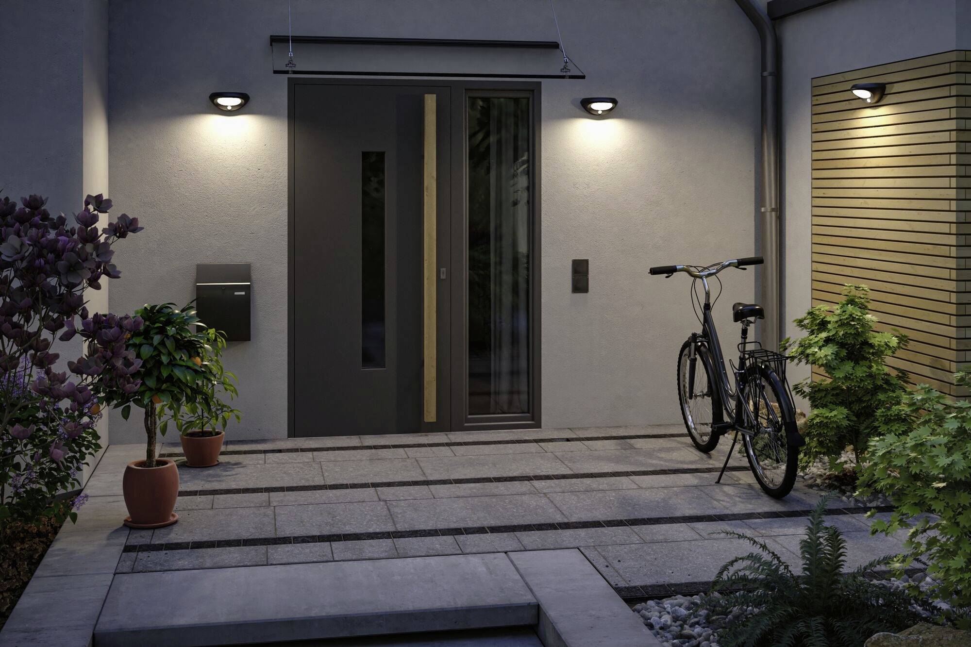 Vstupní prostor domu za soumraku, s moderními dveřmi, osvětlením, jízdním kolem a květináči umístěnými podél kamenné cesty.