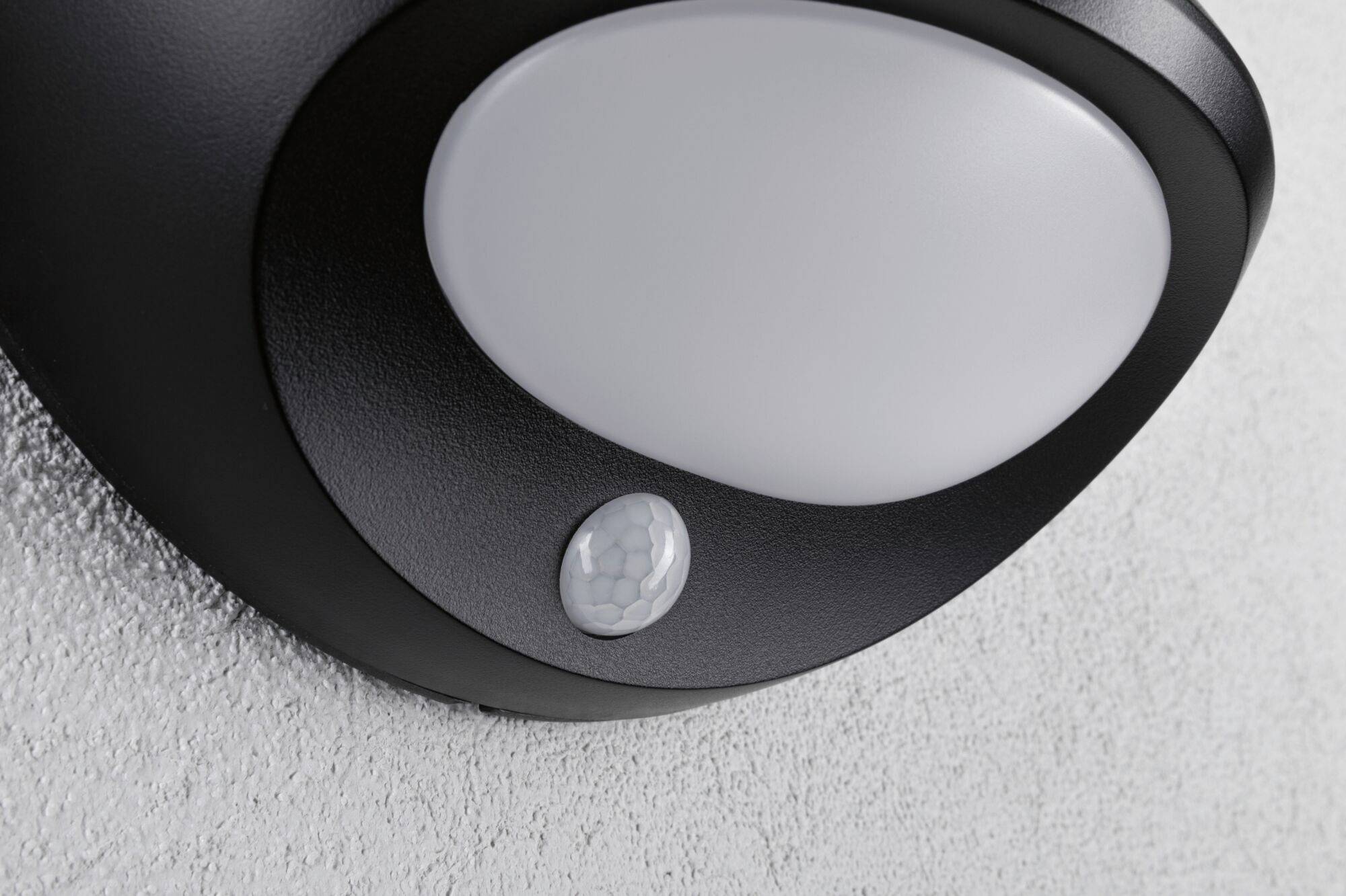 Kulaté černé stropní svítidlo s béžovým senzorem na bílé vymalované stěně.