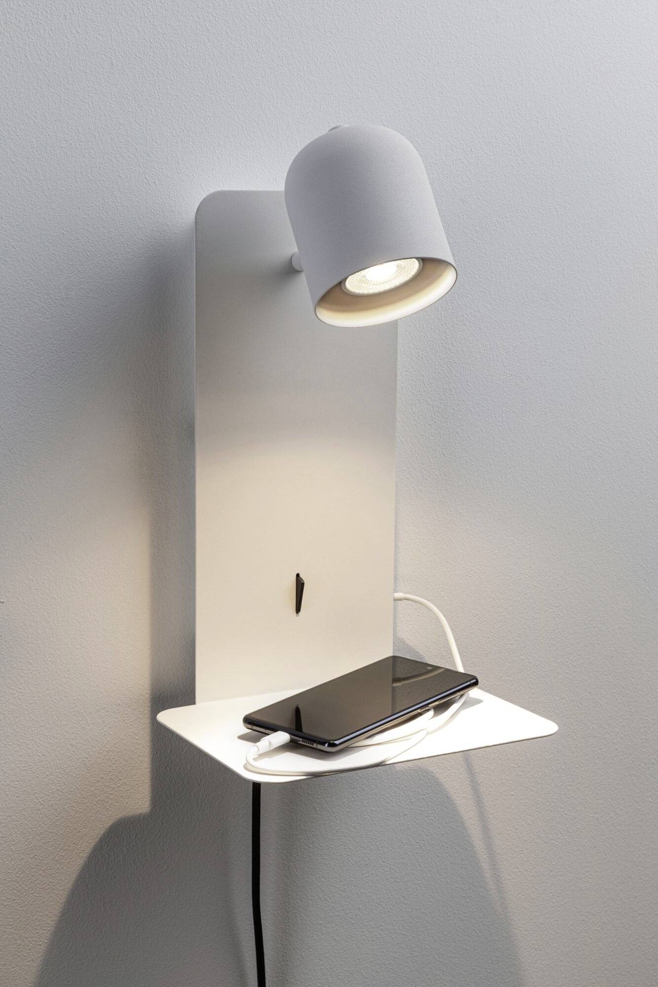Nástěnná lampa s integrovanou policí, na které leží smartphone. Z lampy vychází viditelný nabíjecí kabel.