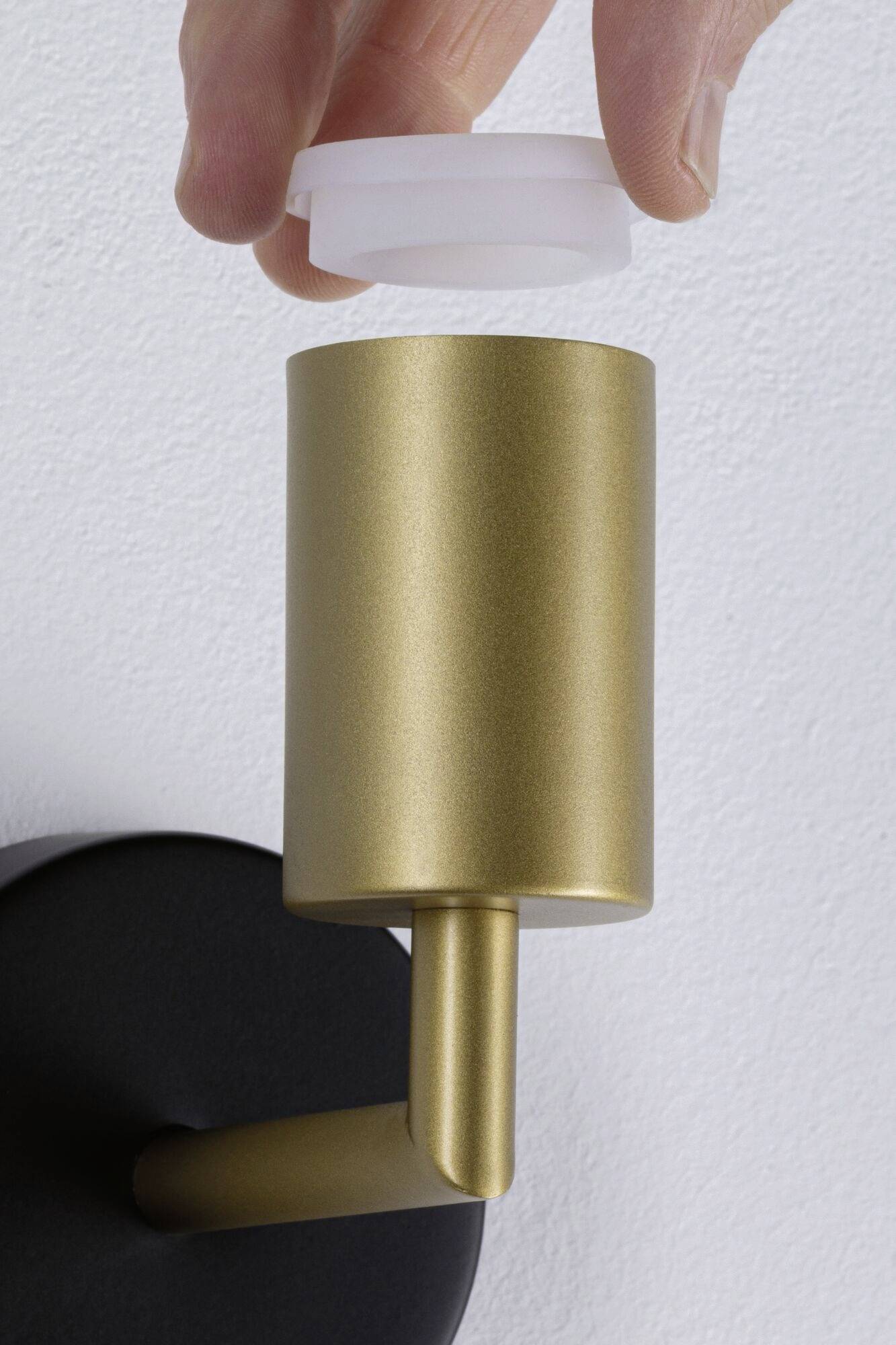Ruka nasazuje bílý kryt na moderní zlatou nástěnnou lampu připevněnou na bílé stěně.