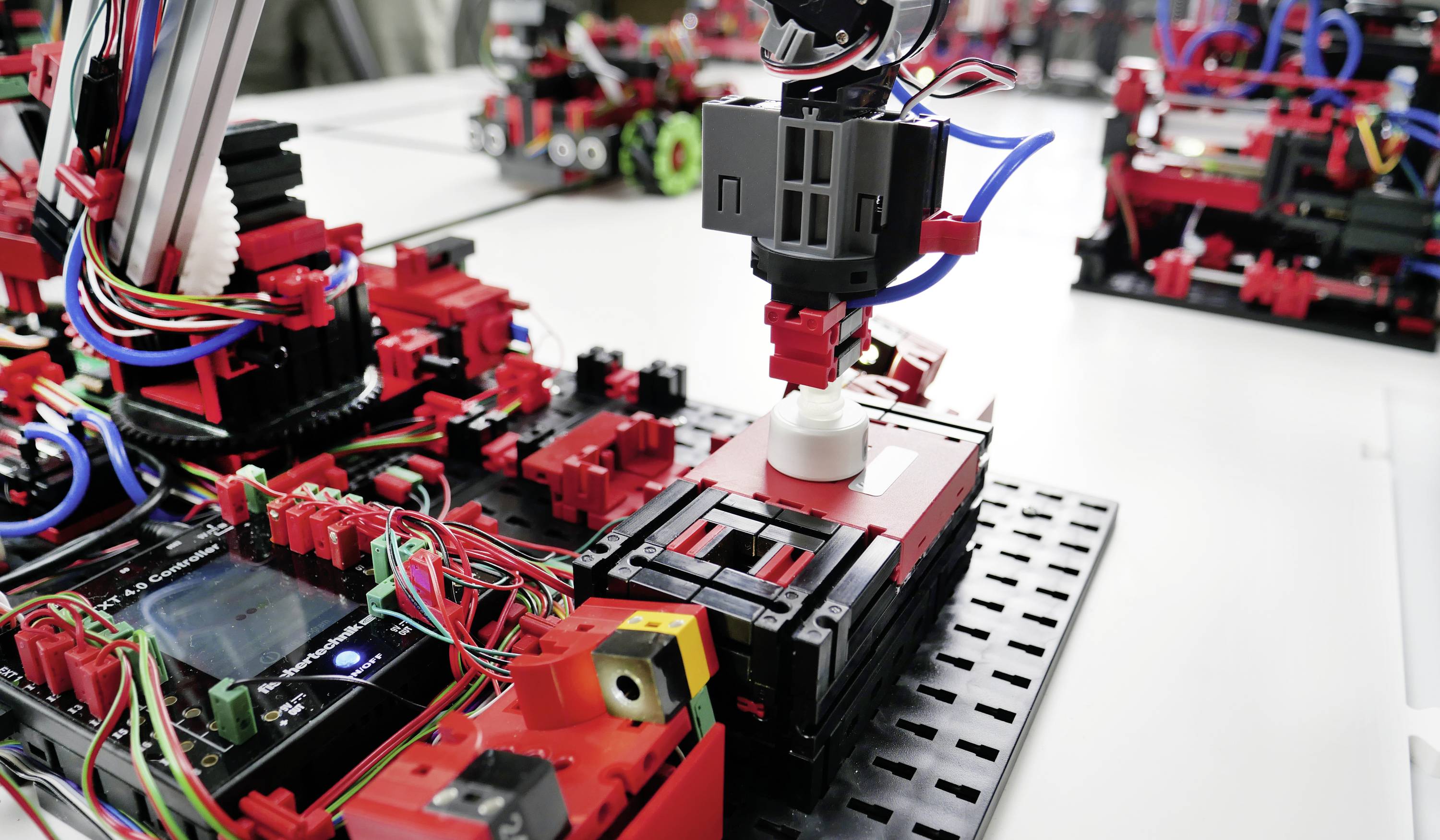 Robotická paže montuje díly modelu na stole plném elektronických součástek a barevných kabelů.