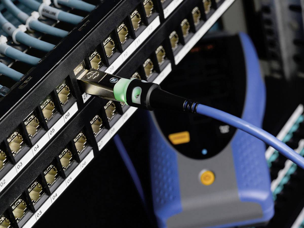 Síťový kabel je zapojen do číslovaného portu serverové skříně. V pozadí je viditelné testovací zařízení.