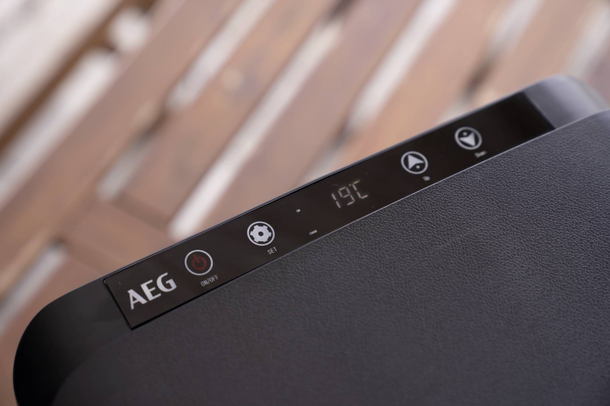 Detail záběr černého elektronického zařízení s digitálním displejem zobrazujícím 19°C. Na zařízení jsou viditelná loga a různé symboly.