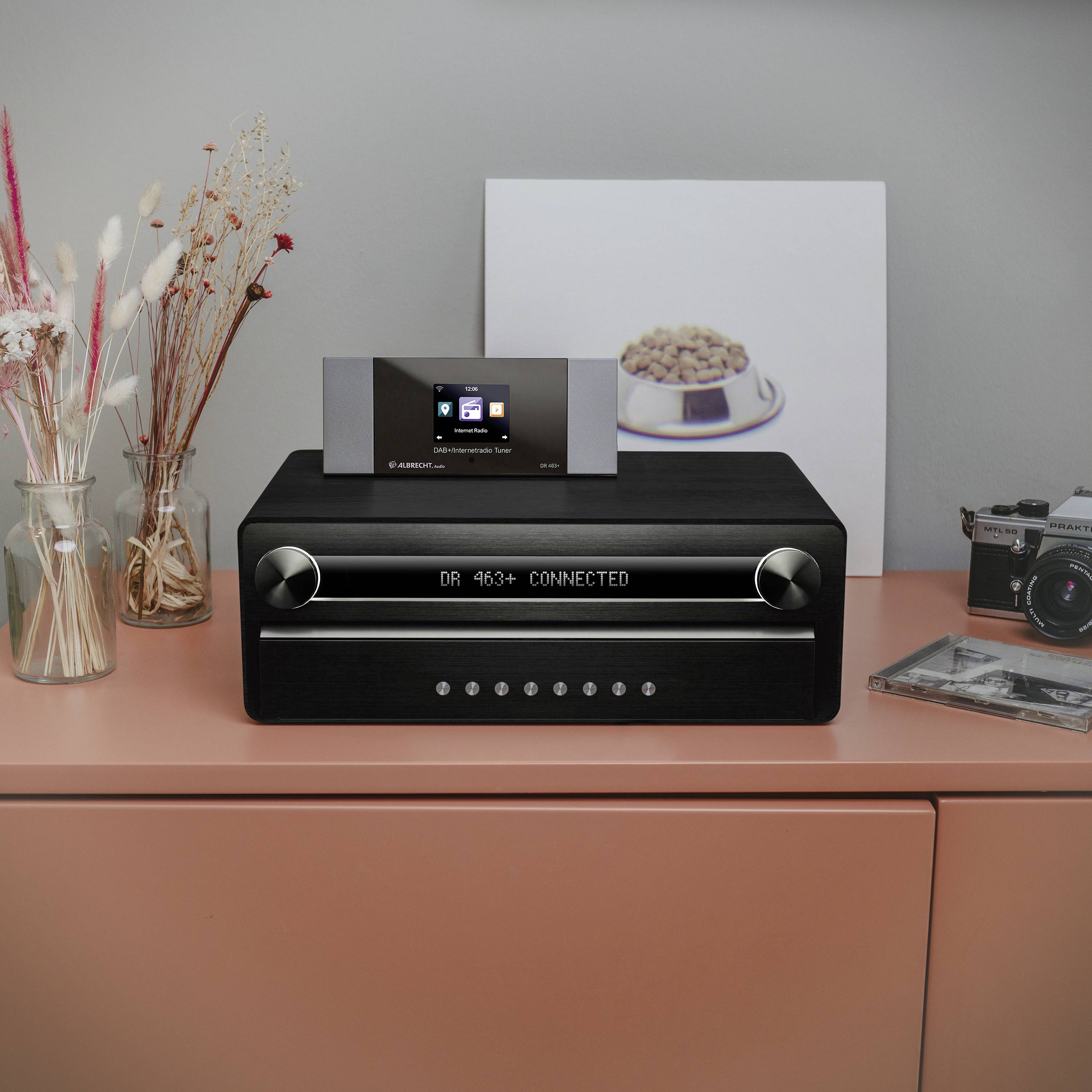 Moderní černé digitální rádio s displejem a dvěma otočnými knoflíky stojí na stole. Vedle něj jsou umístěny fotoaparát a sušené květiny.
