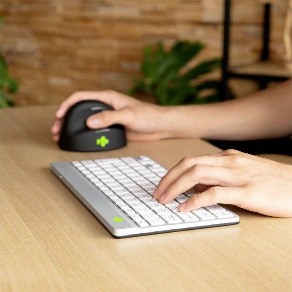 Dvě ruce na psacím stole, jedna drží ergonomickou myš, druhá píše na ploché klávesnici se zelenými akcentními klávesami.
