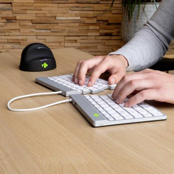 Osoba píše na sdílené ergonomické klávesnici položené na dřevěném stole. V pozadí je myš a pokojová rostlina.