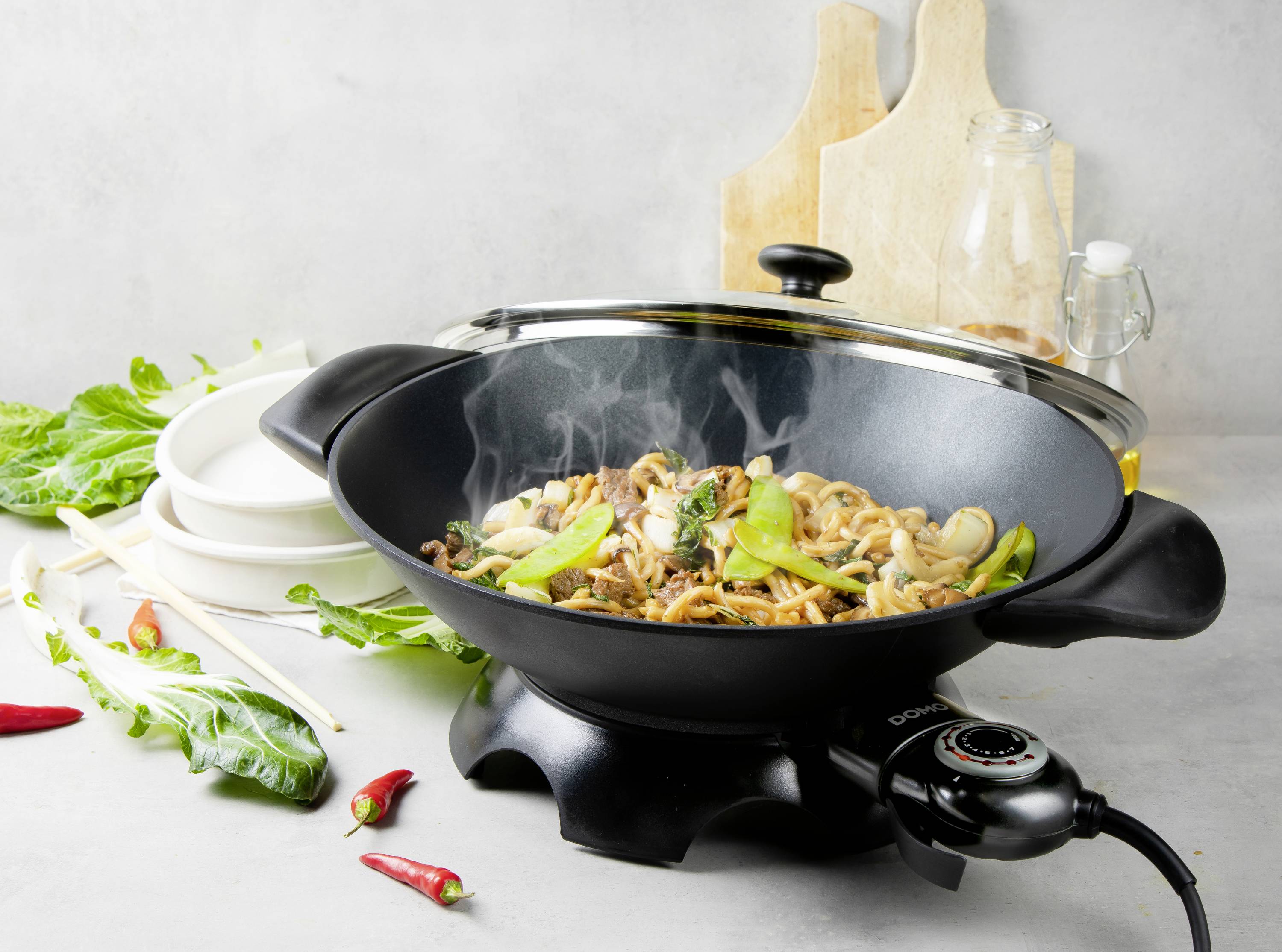Wok s párou stoupající z asijské zeleninové pánve, složené z nudlí, zelené zeleniny a hub. V pozadí koření a talíře.