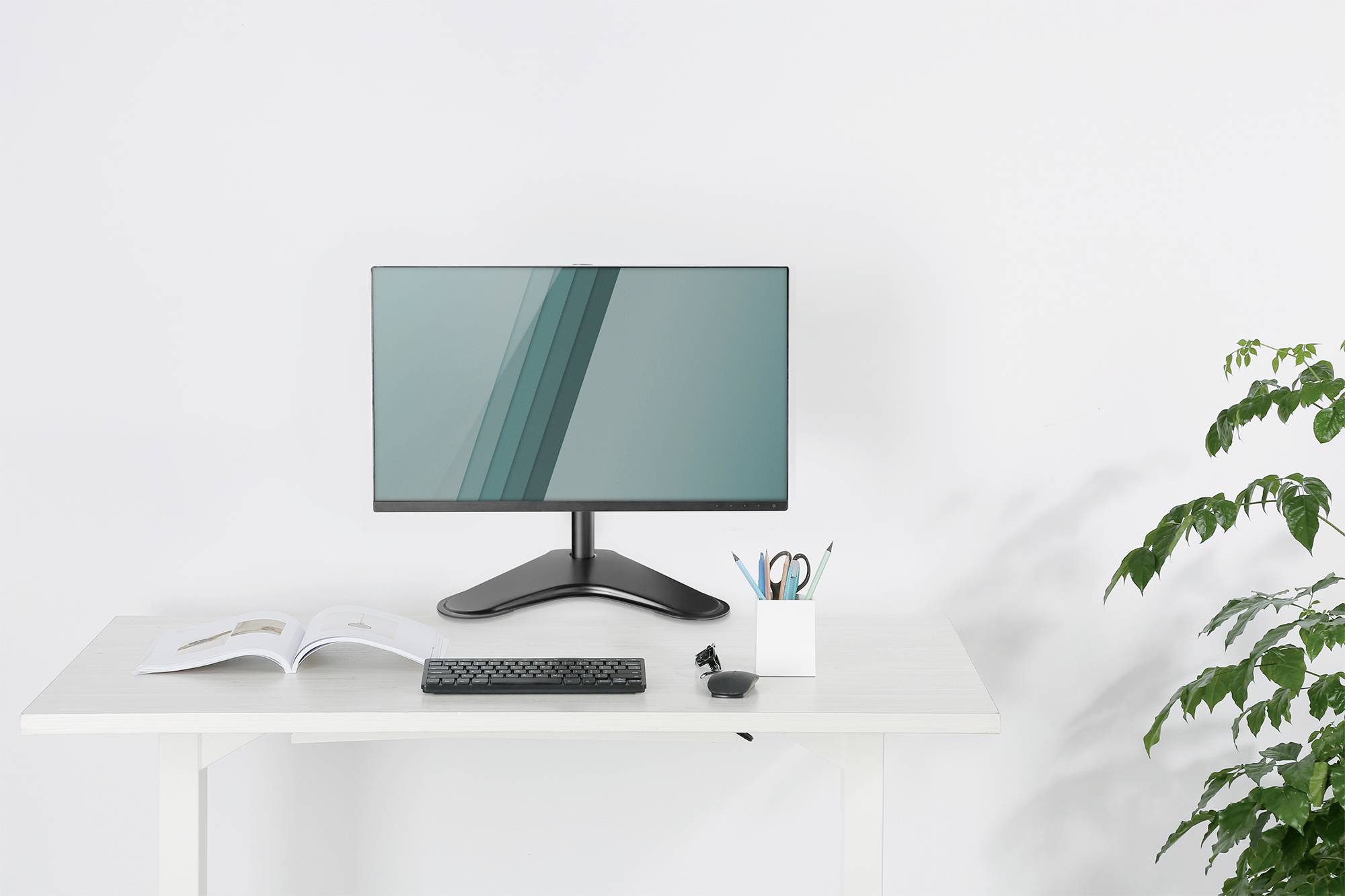 Moderní počítačový monitor na bílém psacím stole. Vedle něj jsou klávesnice, otevřená kniha, stojánek na tužky a rostlina.
