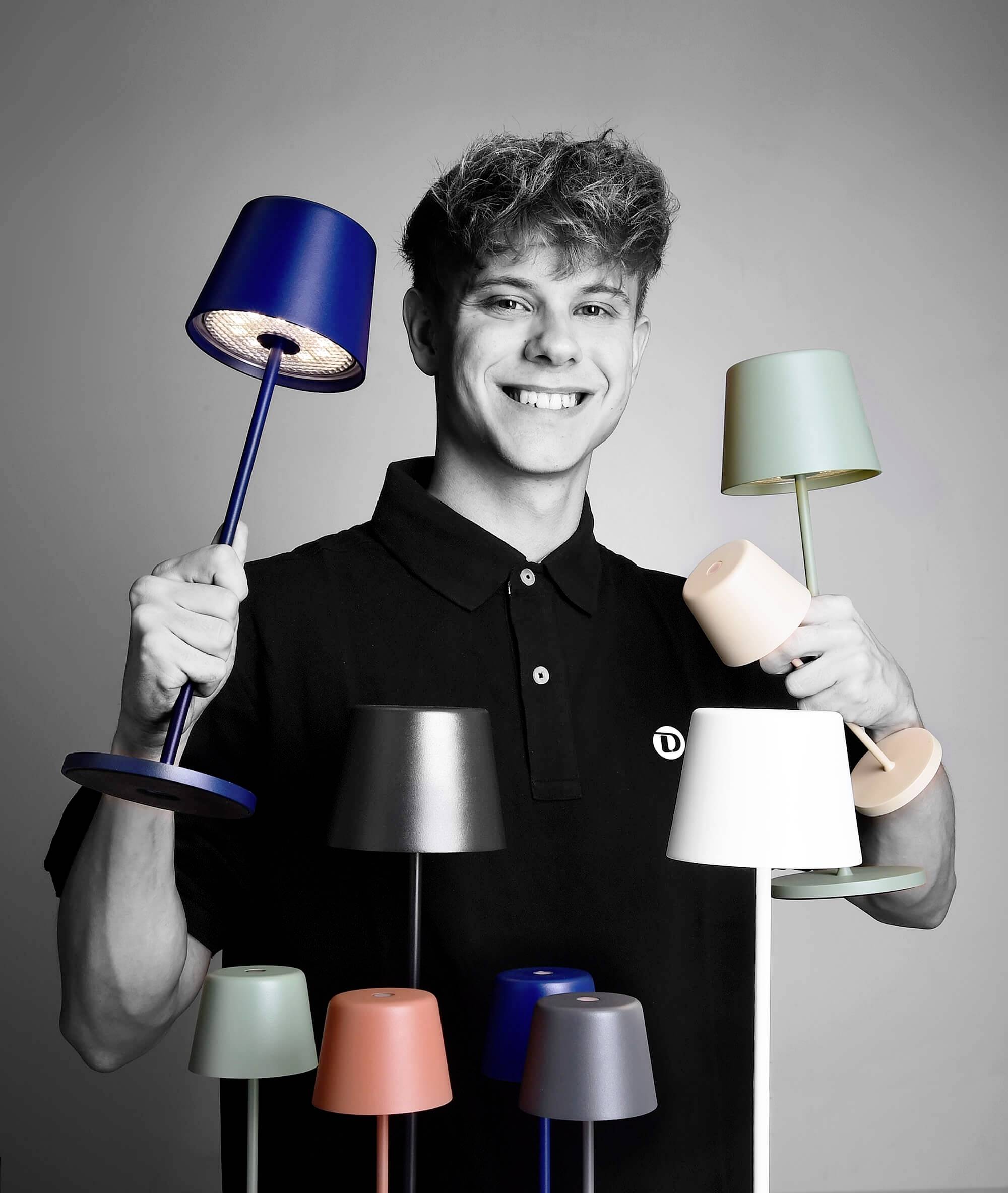 Usmívající se muž drží v rukou několik barevných stolních lamp, obklopen podobně vyhlížejícími lampami.