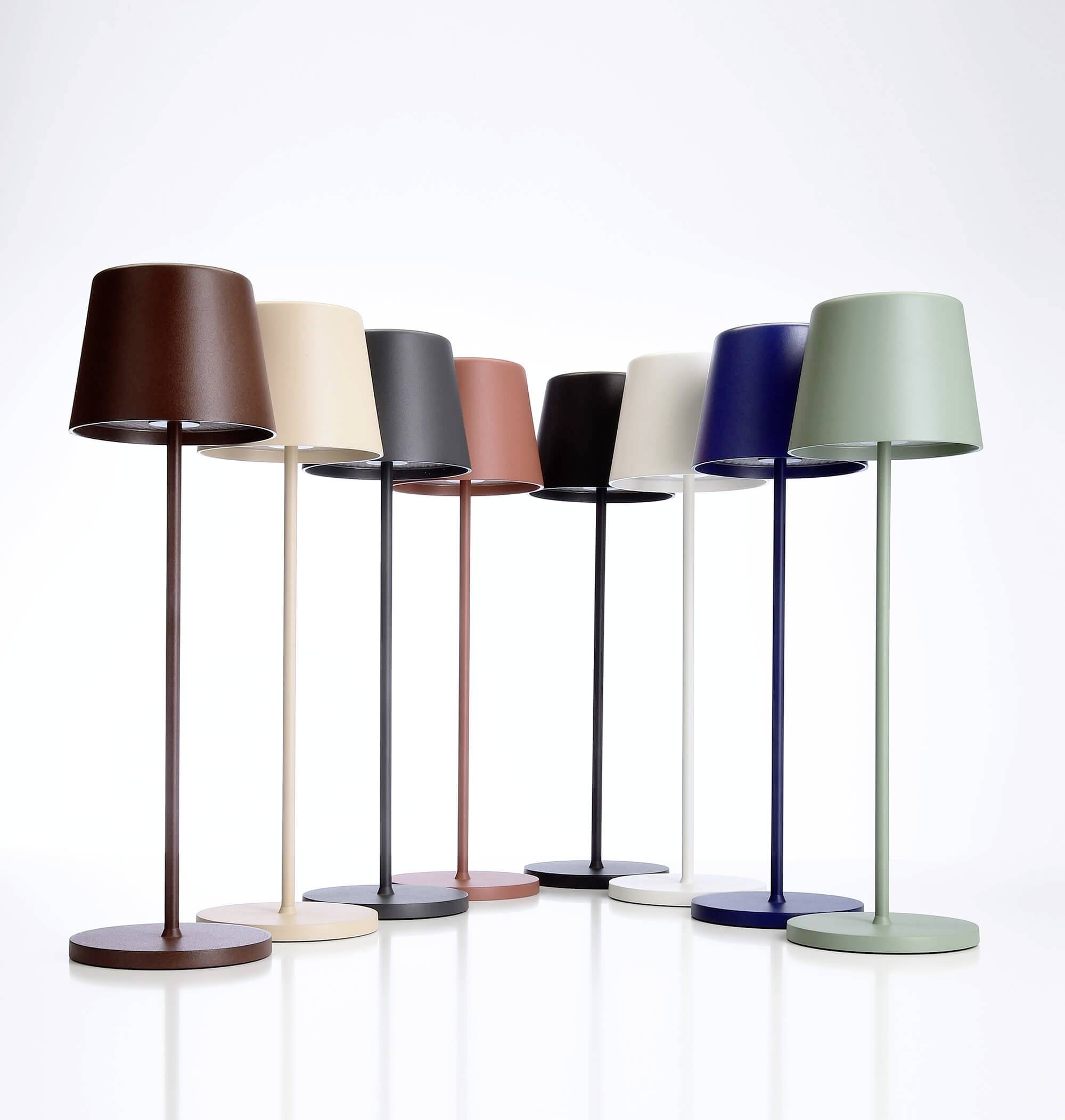 Osm barevných stolních lamp v řadě na bílém pozadí, v odstínech od hnědé, krémové, růžové, černé, modré až po zelenou.