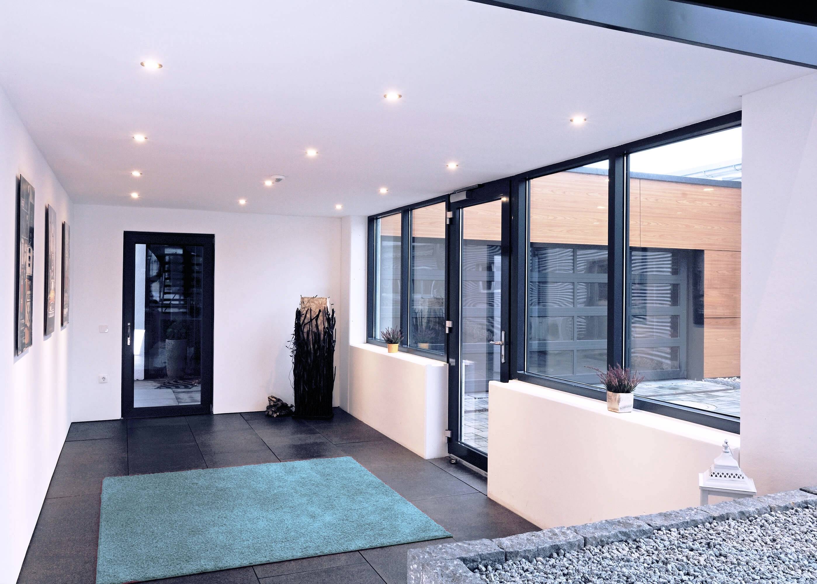 Moderní chodba s velkými okny a jasným osvětlením, výhled do vnitřního dvora s kamenným povrchem; jednoduchá výzdoba.