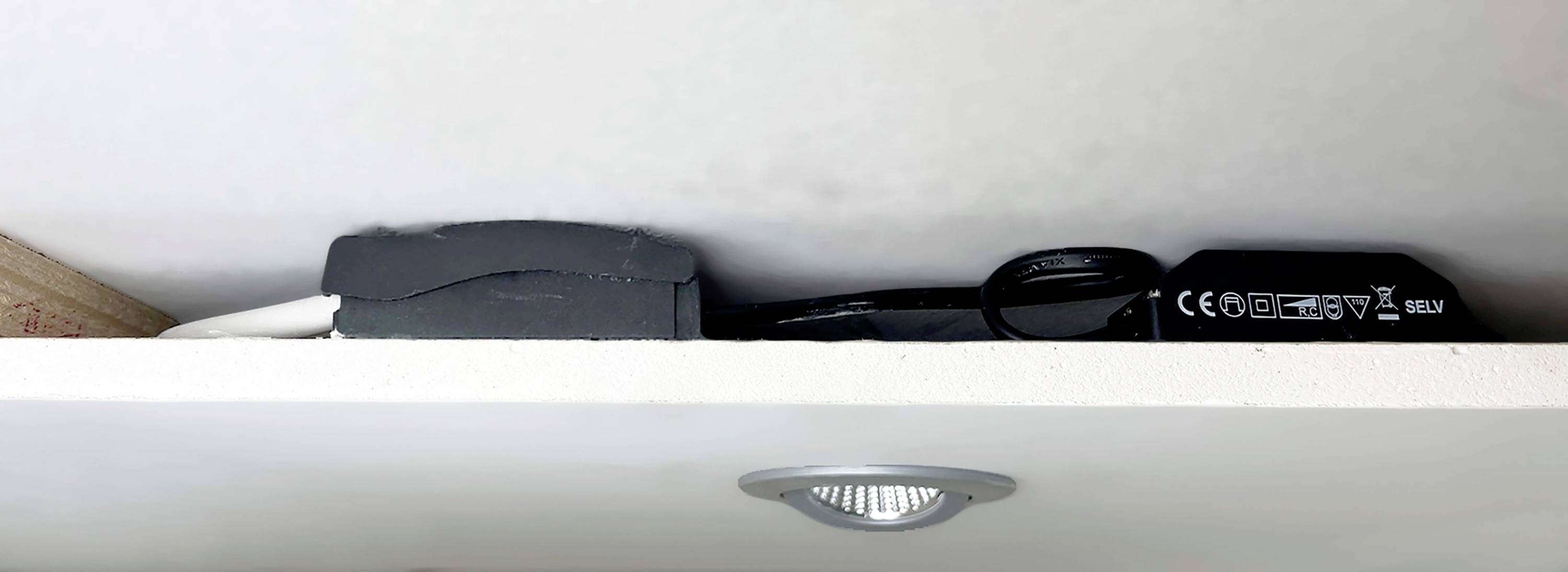 Černé elektronické zařízení s kabely leží na bílé polici. Pod ním je viditelná kulatá stropní lampa.