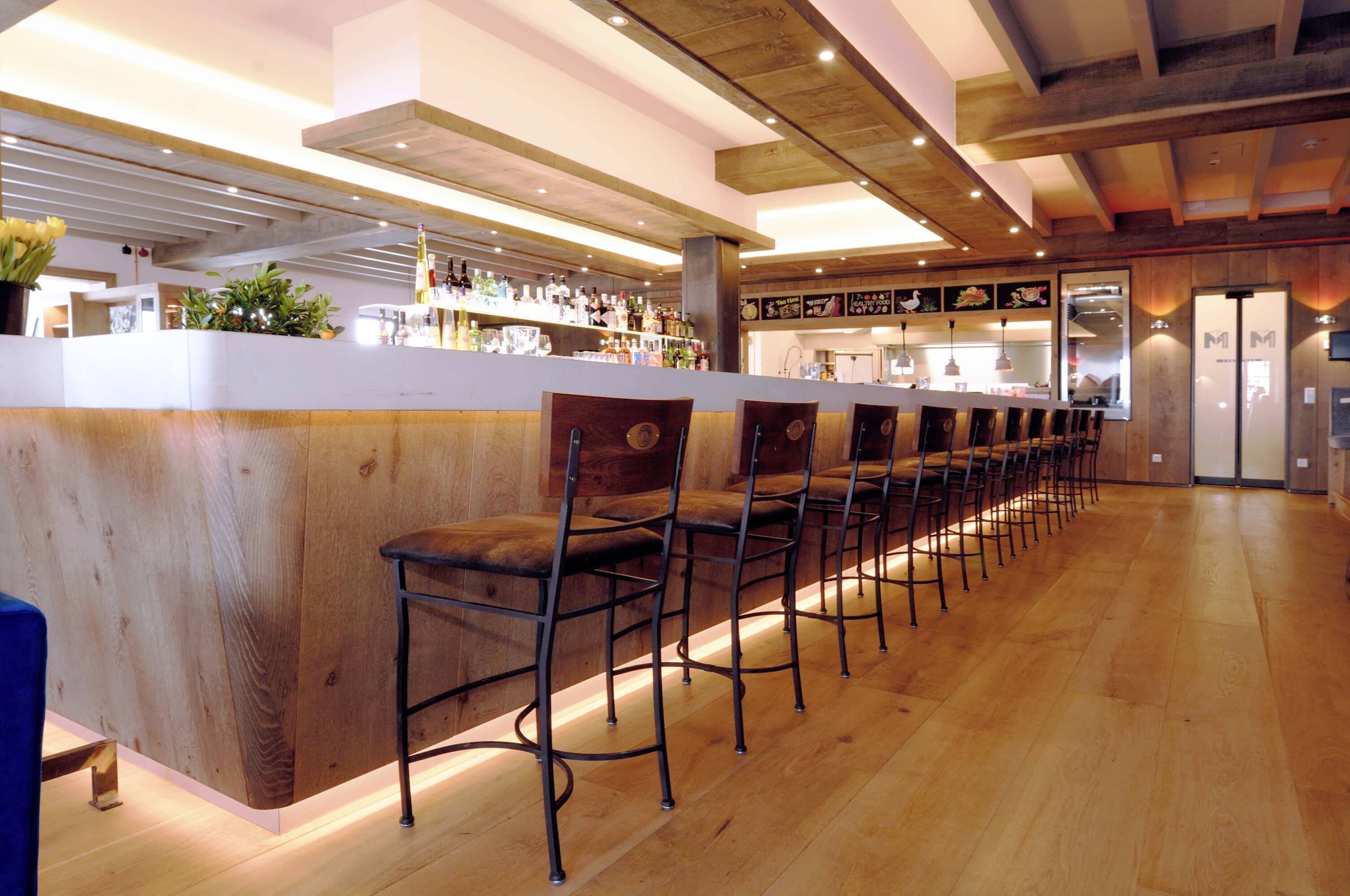 Moderní restaurace s dlouhým dřevěným barem a vysokými stoličkami, útulným osvětlením a rostlinami vytváří příjemnou atmosféru.