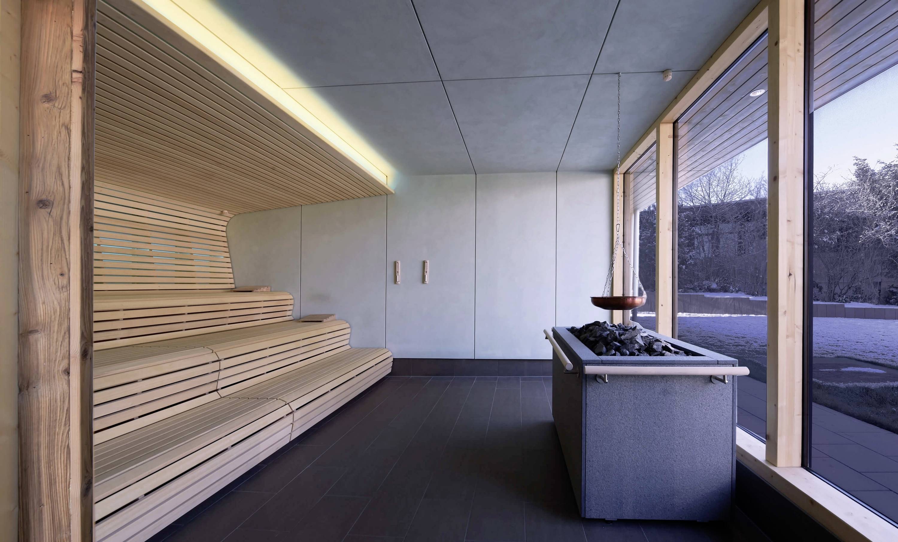 Moderní sauna s dřevěnými sedadly a pecí na pravé straně. Velká okna nabízejí výhled na zasněnou krajinu.