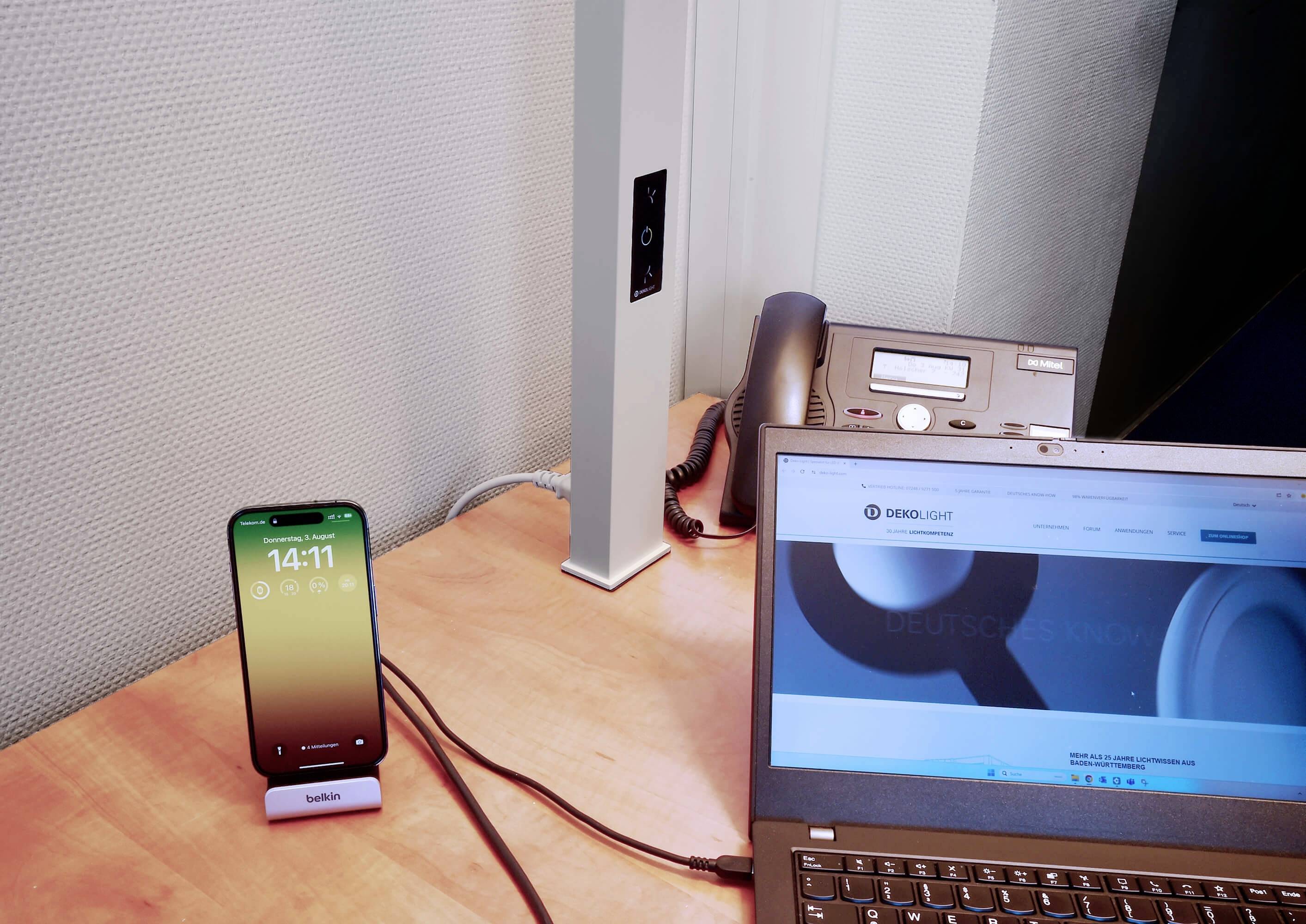 Mobilní telefon na psacím stole zobrazuje čas. Vedle něj je laptop s otevřenou webovou stránkou a stolní telefon.