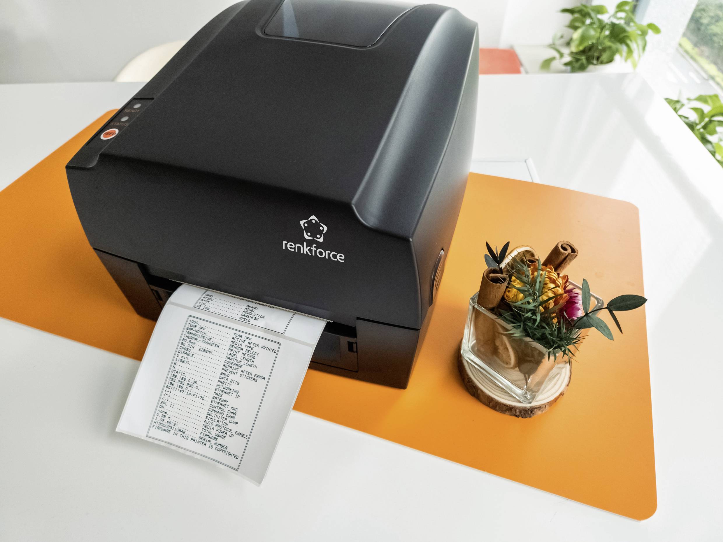 Termální tiskárna na stole tiskne účtenku. Vedle ní stojí rostliny na oranžovém prostírání.