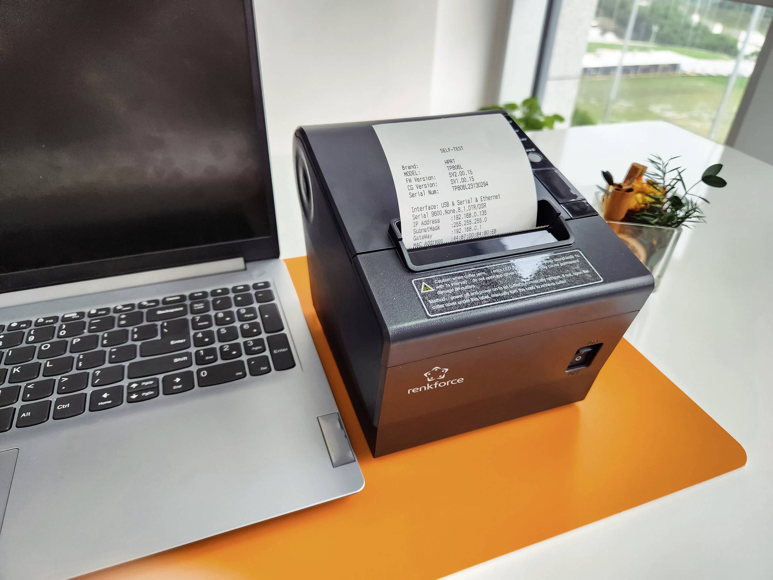 Notebook vedle tiskárny, která tiskne účtenku. Tiskárna stojí na oranžovém stole, poblíž jsou rostliny.
