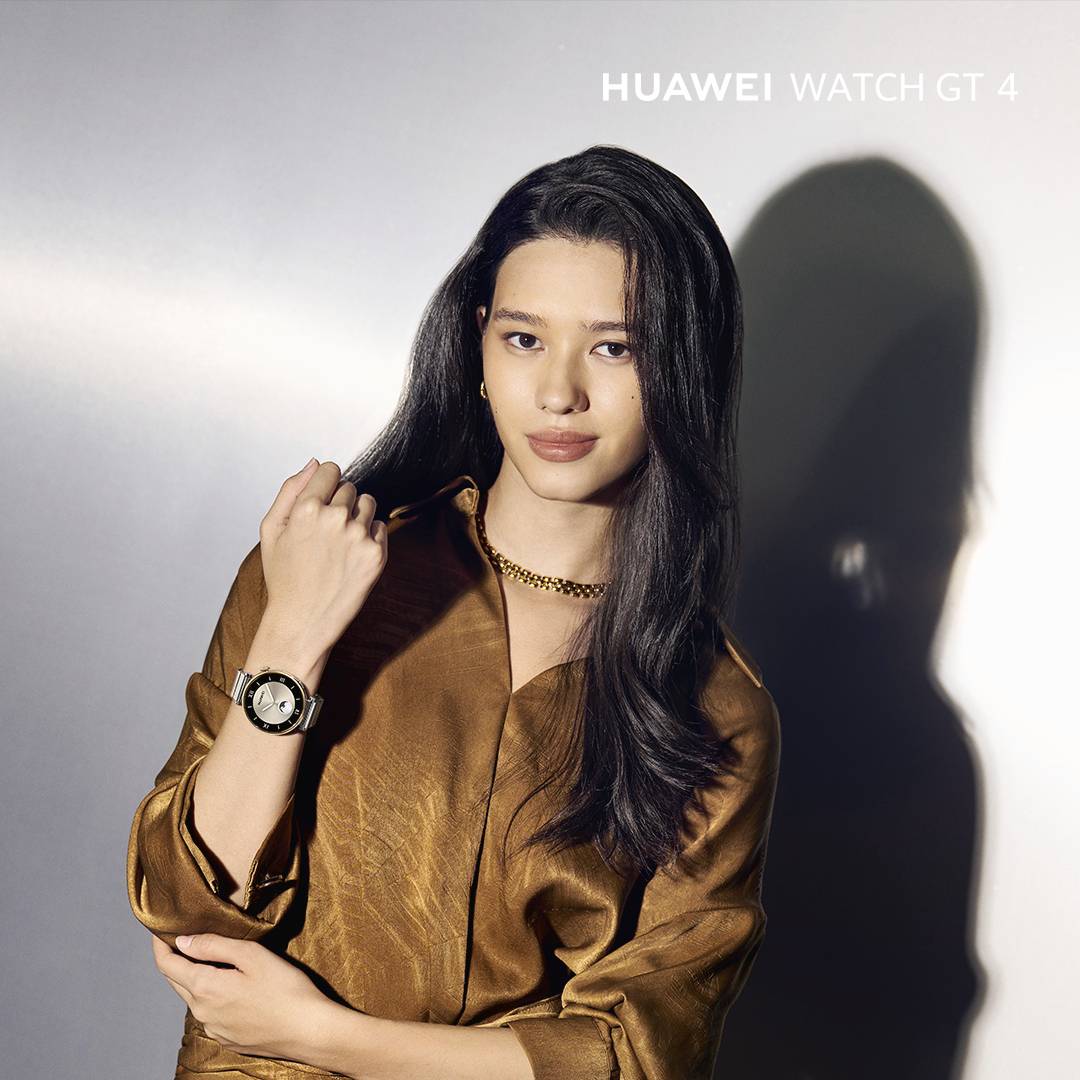 Žena ve zlatém tričku nosí náramkové hodinky, model 'Huawei Watch GT 4'. Hodinky mají kulatý ciferník s viditelným řemínkem.