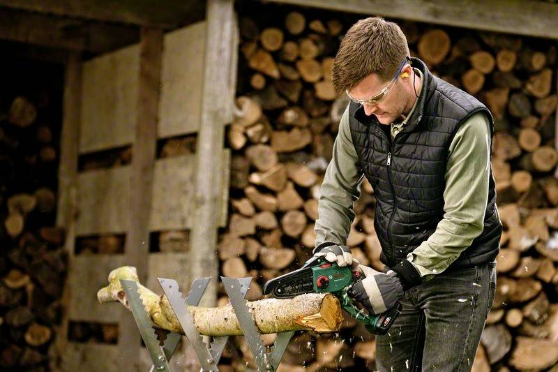 Muž v bezpečnostních brýlích a chráničích sluchu rozřezává motorovou pilou kmen dřeva na pile se svěrákem. V pozadí je vidět naskládané palivové dřevo.