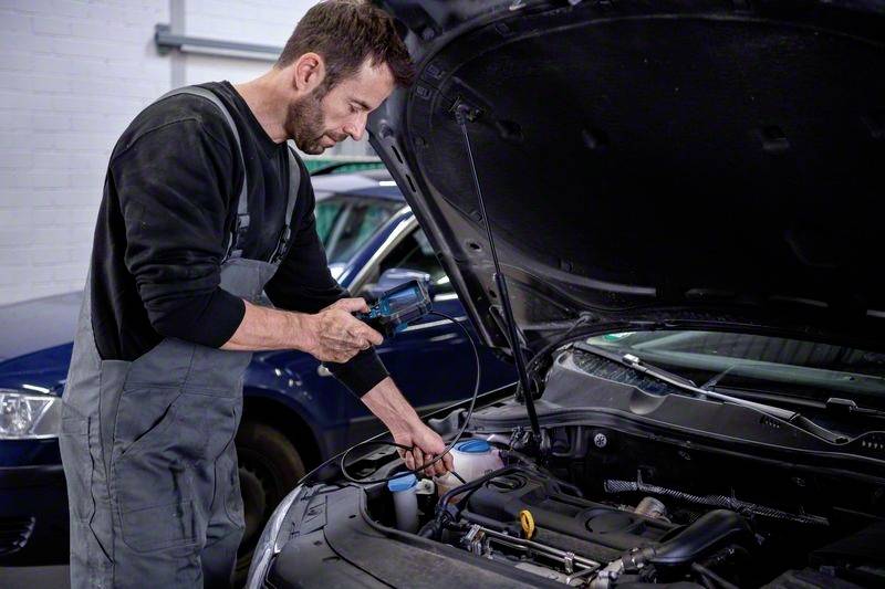 Mechanik kontroluje otevřený motorový prostor automobilu v autoservisu. V pozadí stojí další automobil.