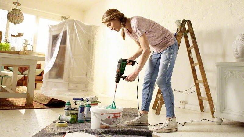 Žena míchá barvu v renovovaném obývacím pokoji. Kolem ní jsou rozmístěné krycí plachty, malířské potřeby a žebřík.