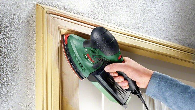 Osoba používá elektrickou delta brusku k broušení okenního rámu. Přístroj má zelený kryt a červený brusný nástavec.