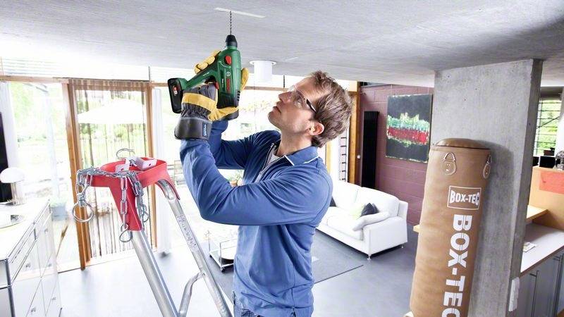 Muž na žebříku vrtá do stropu obývacího pokoje pomocí elektrické vrtačky. V pozadí je boxovací pytel.