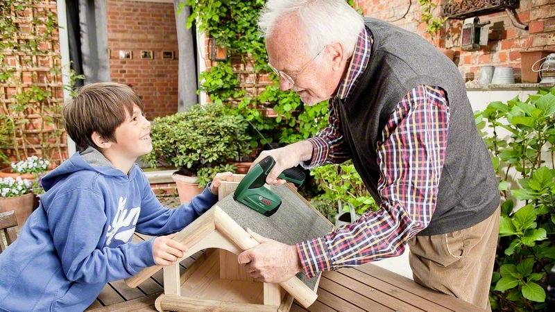 Starší muž a chlapec společně staví dřevěnou ptačí budku na zahradě. Muž drží nástroj, zatímco chlapec přihlíží.