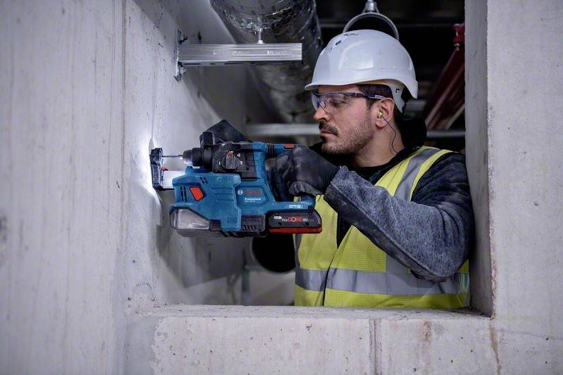 Dělník v bezpečnostním oblečení vrtá modrou vrtačkou díru do betonové zdi.